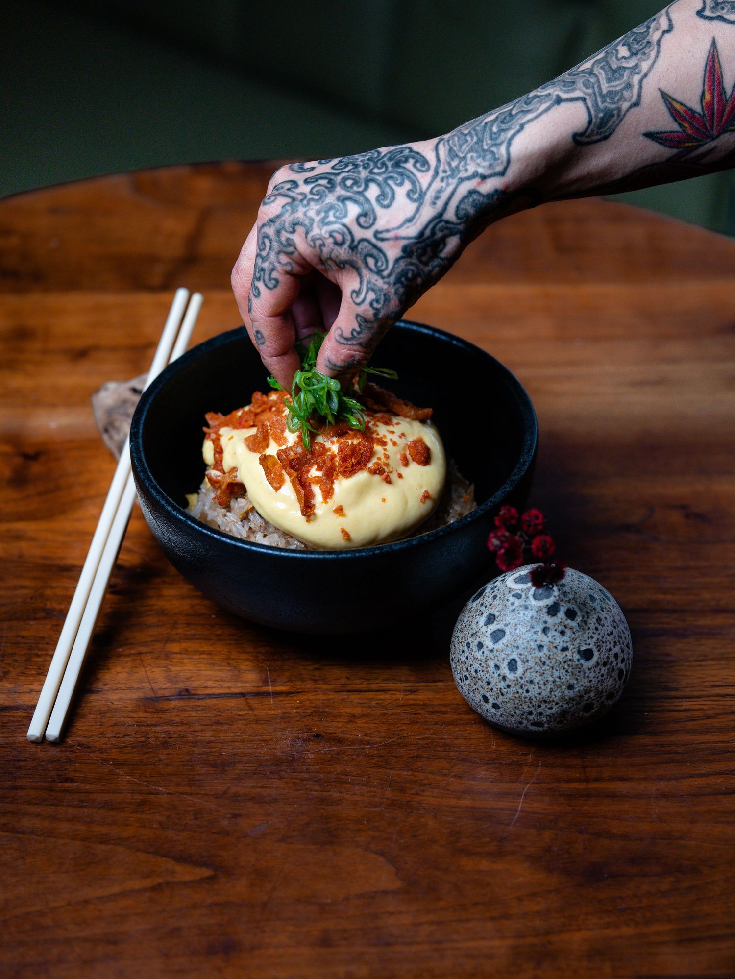 Tattooed hand garnishing a black bowl of rice with creamy sauce and toppings, chopsticks on wooden table.