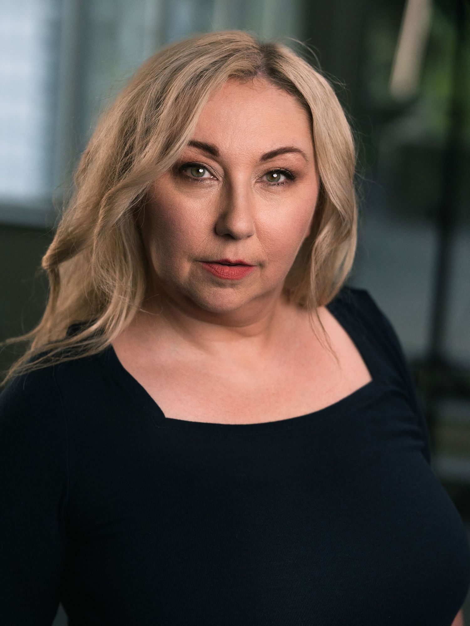 Blonde woman in black top poses confidently for a professional headshot portrait indoors.