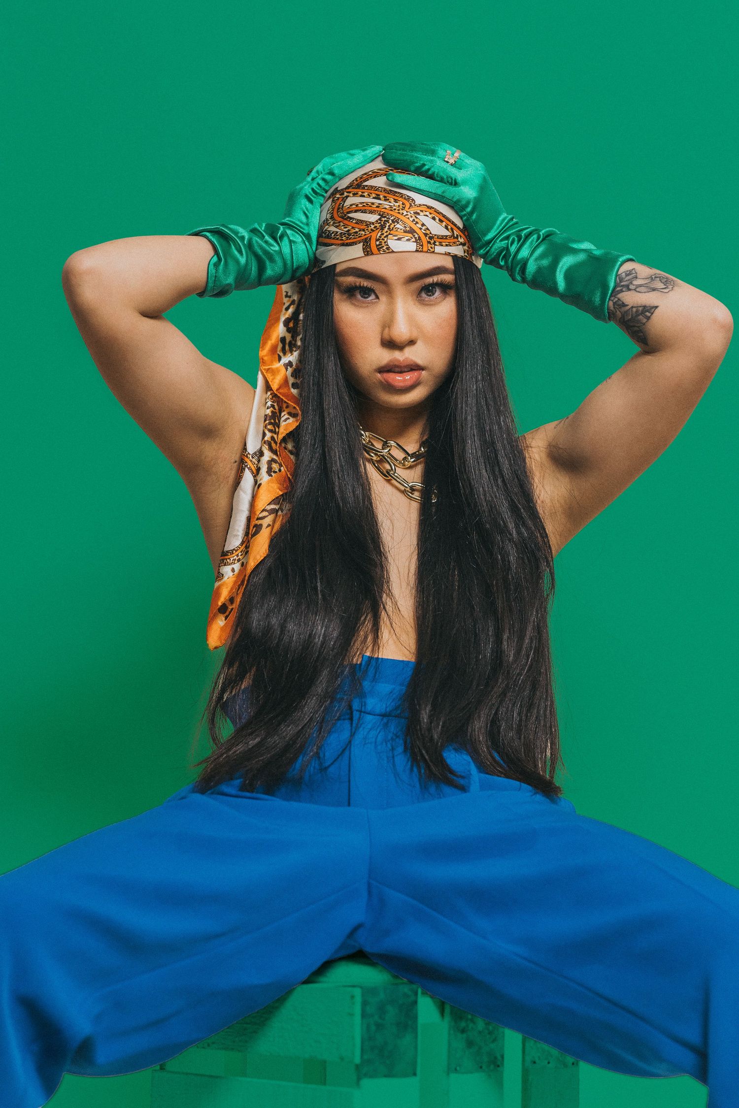 Woman in blue jumpsuit and green gloves poses confidently against a green background wearing a patterned headscarf.