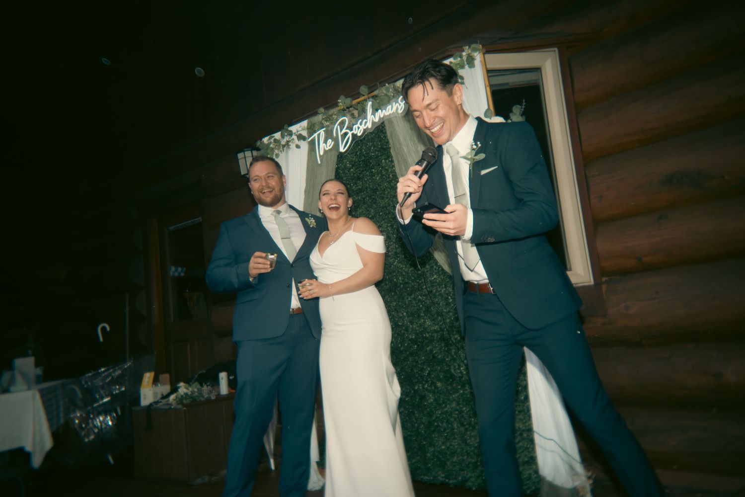 Bride and groom celebrate with a guest at wedding reception in rustic log cabin venue.