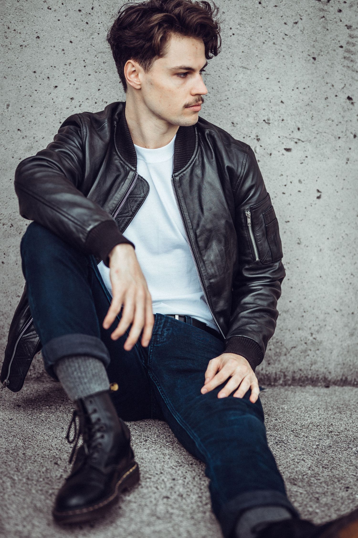 Young man in black leather jacket, white tee, and jeans sitting against concrete wall wearing boots. Brand content photography