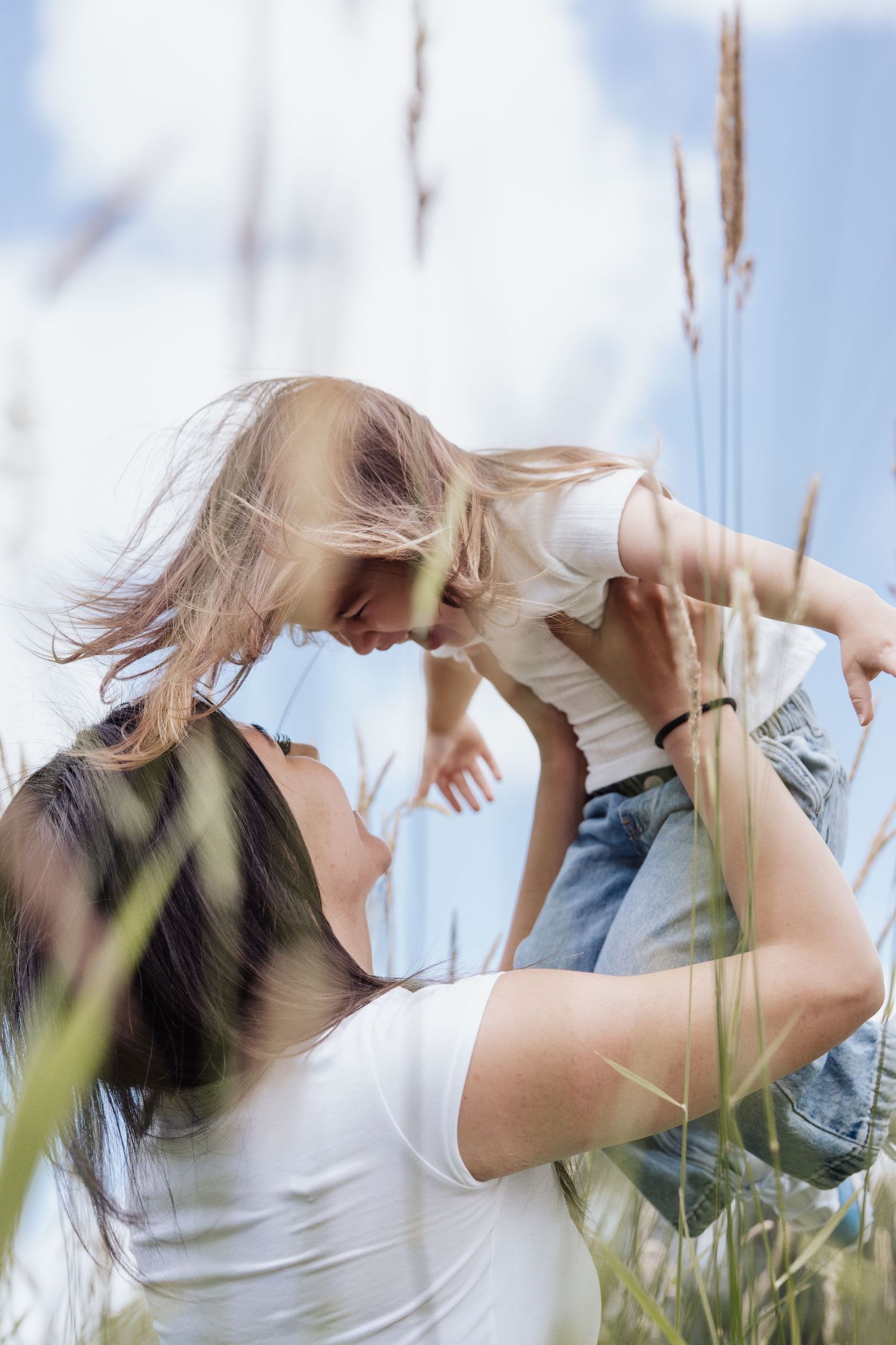 Mother lifting her young daughter in a field of tall grass under a blue sky, sharing a joyful moment.