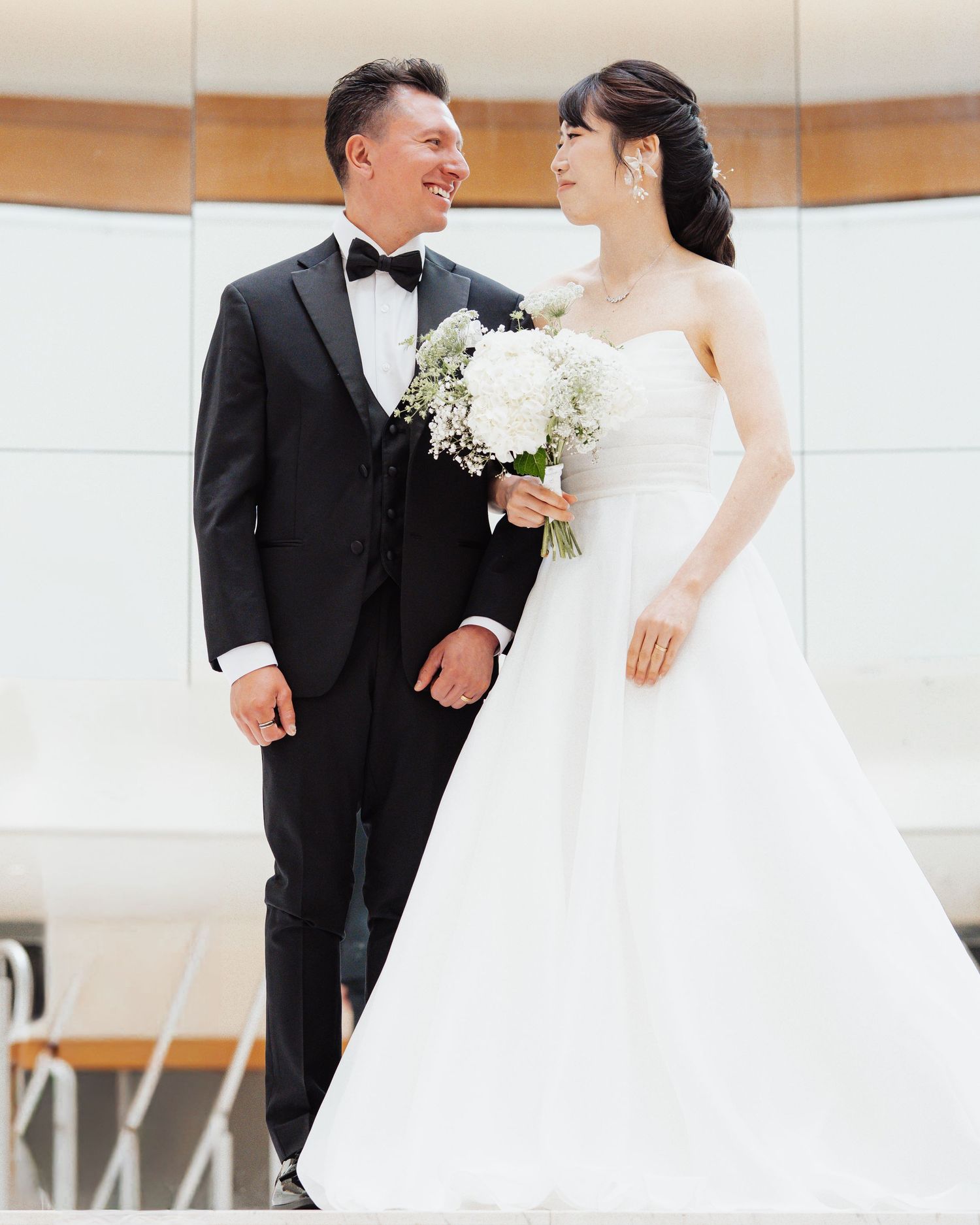 A couple in wedding attire smiles at each other, groom in black tuxedo, bride in white strapless gown holding white bouquet.