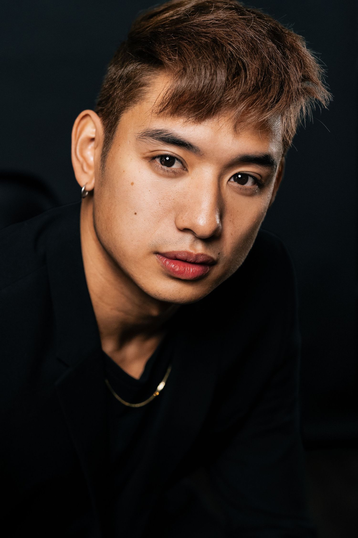 Young Asian man with brown hair, small earring, and gold necklace poses against dark background.