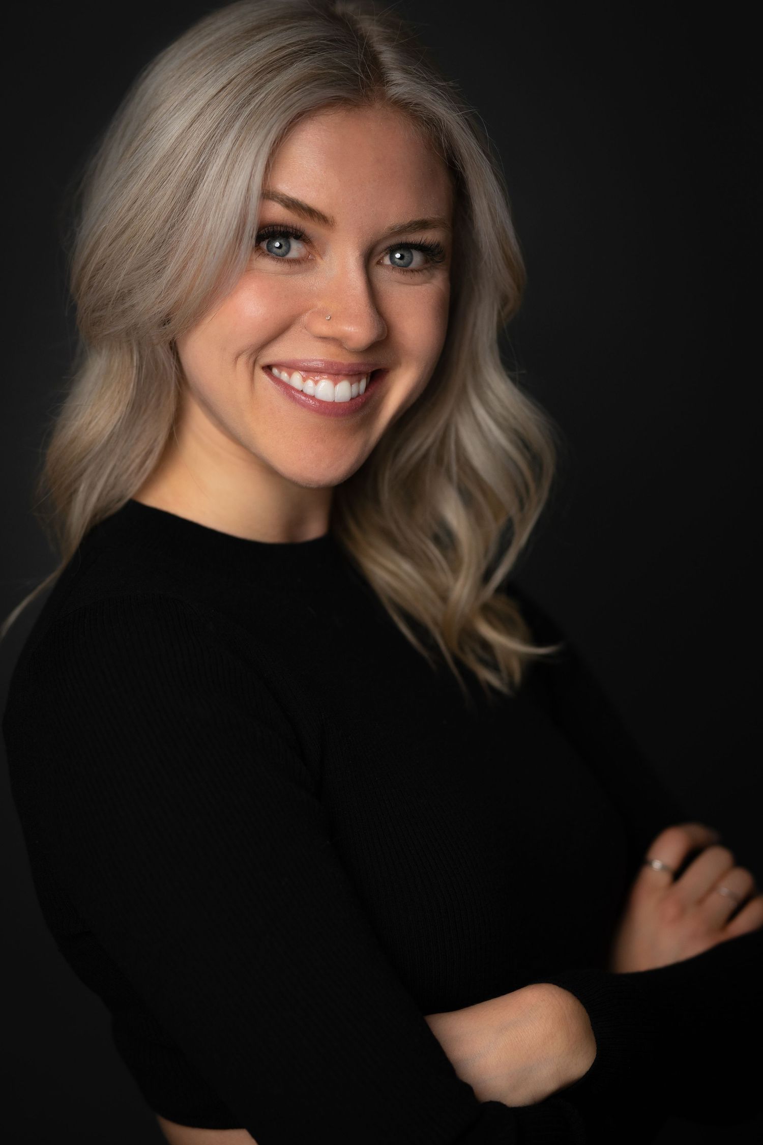 Smiling blonde woman in black top with arms crossed against dark background, professional headshot photo.