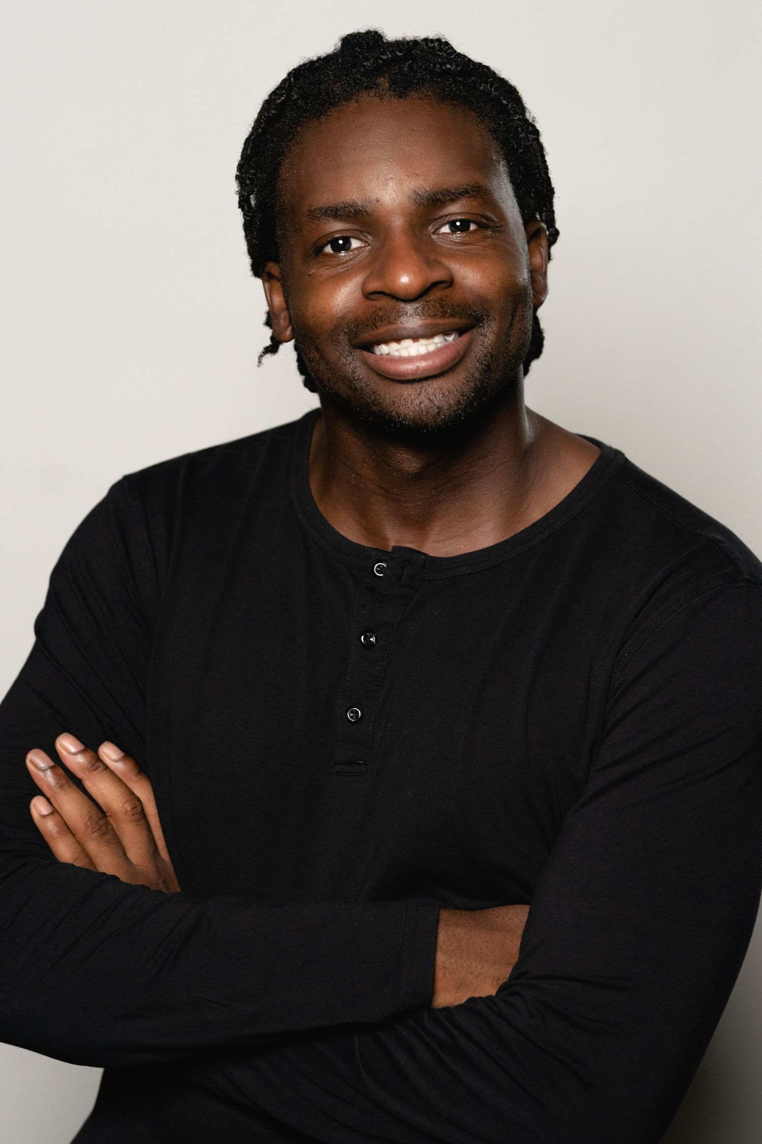Smiling Black man with braids wearing a black henley shirt, arms crossed, posing against a light gray background.