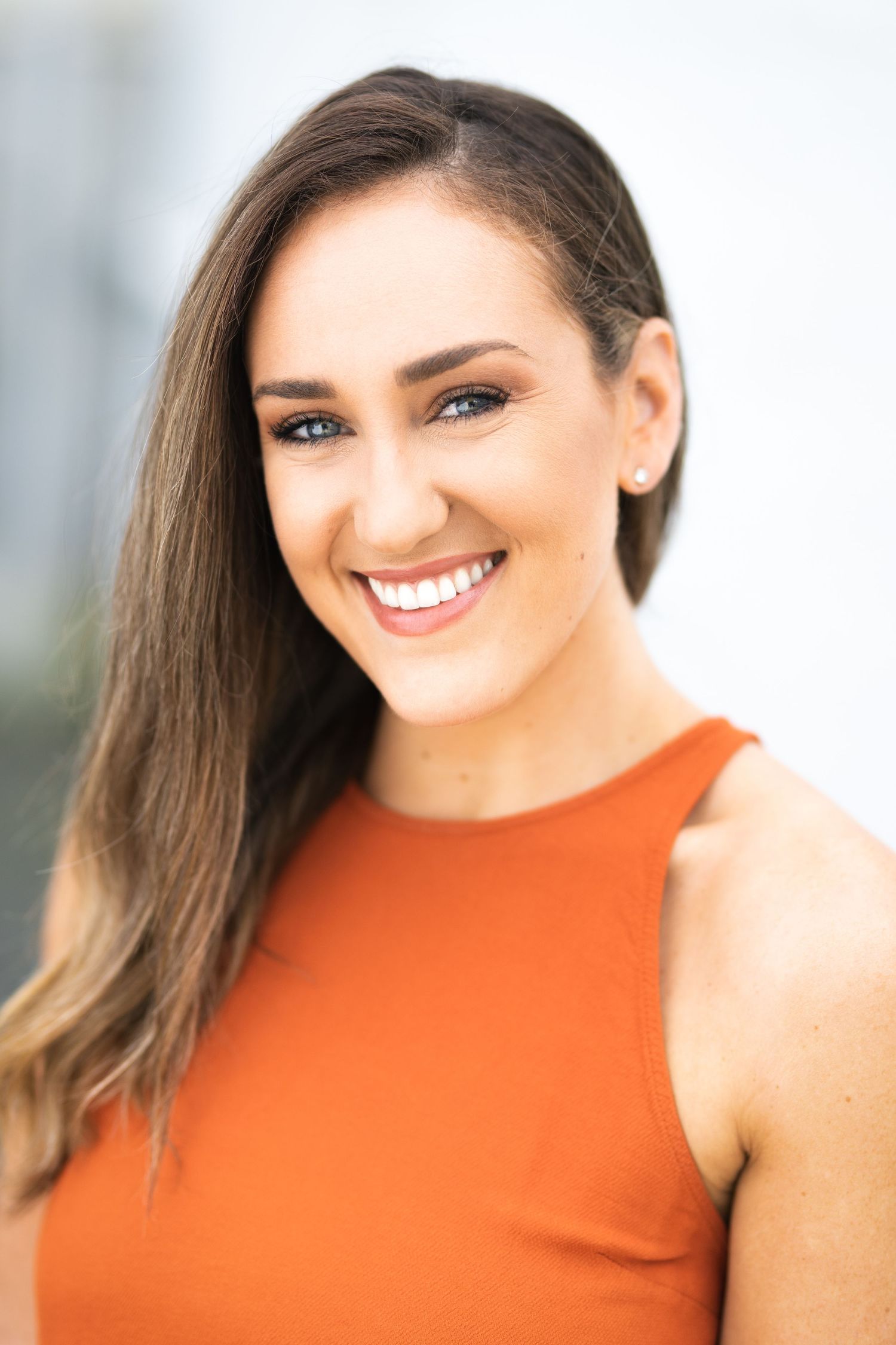 Smiling young woman with long brown hair wearing an orange sleeveless top, posing outdoors in a professional headshot.