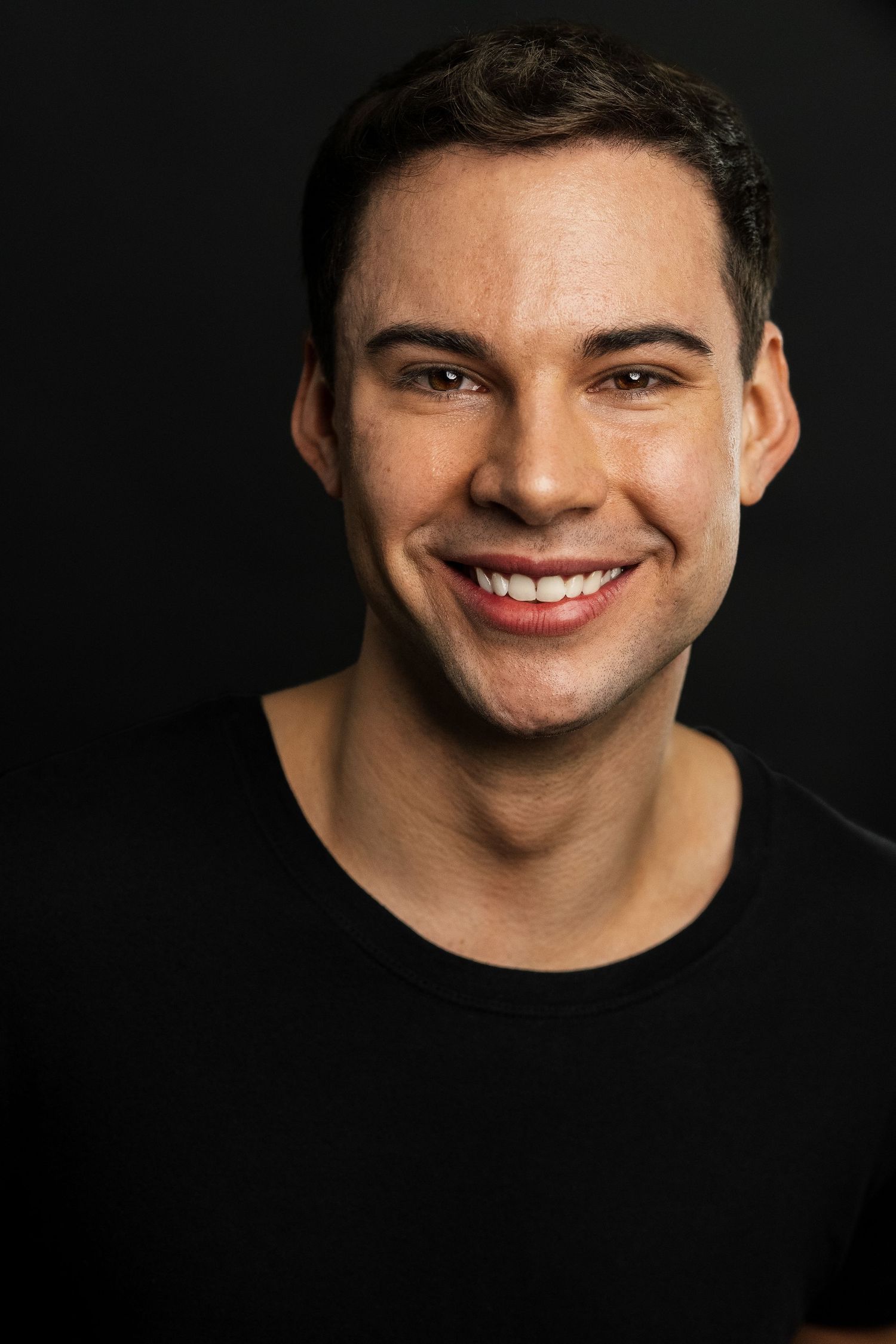 Smiling young man with short dark hair wearing a black t-shirt against a dark background.