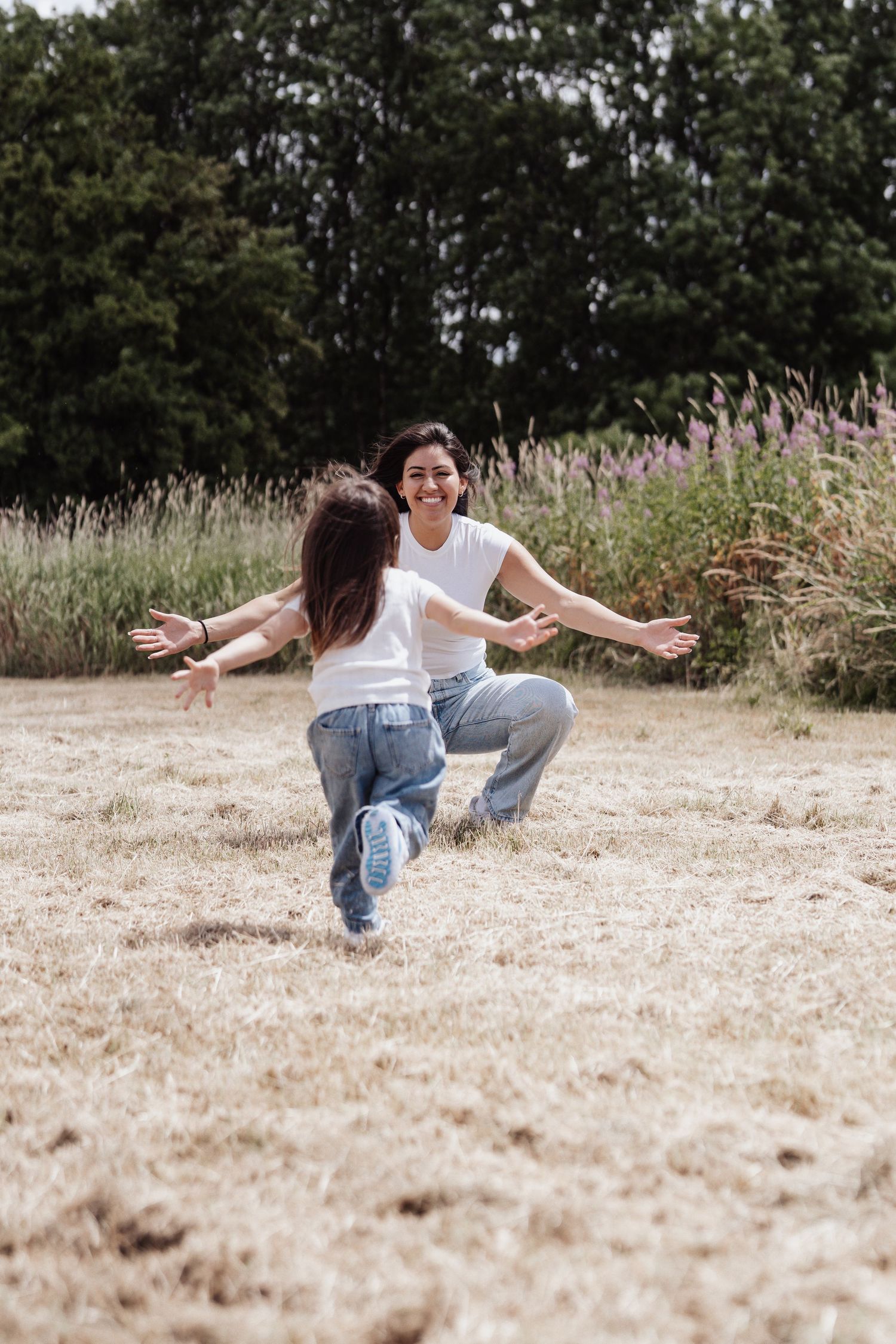 A joyful mother crouches with open arms as her young daughter runs toward her in a sunny field.