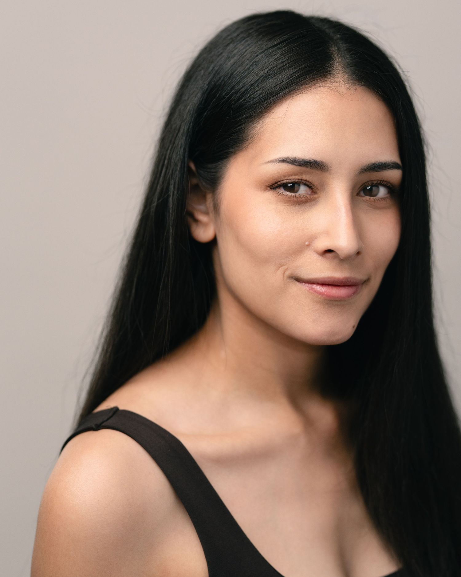 Young woman with long black hair smiling softly, wearing a black top against a neutral background.