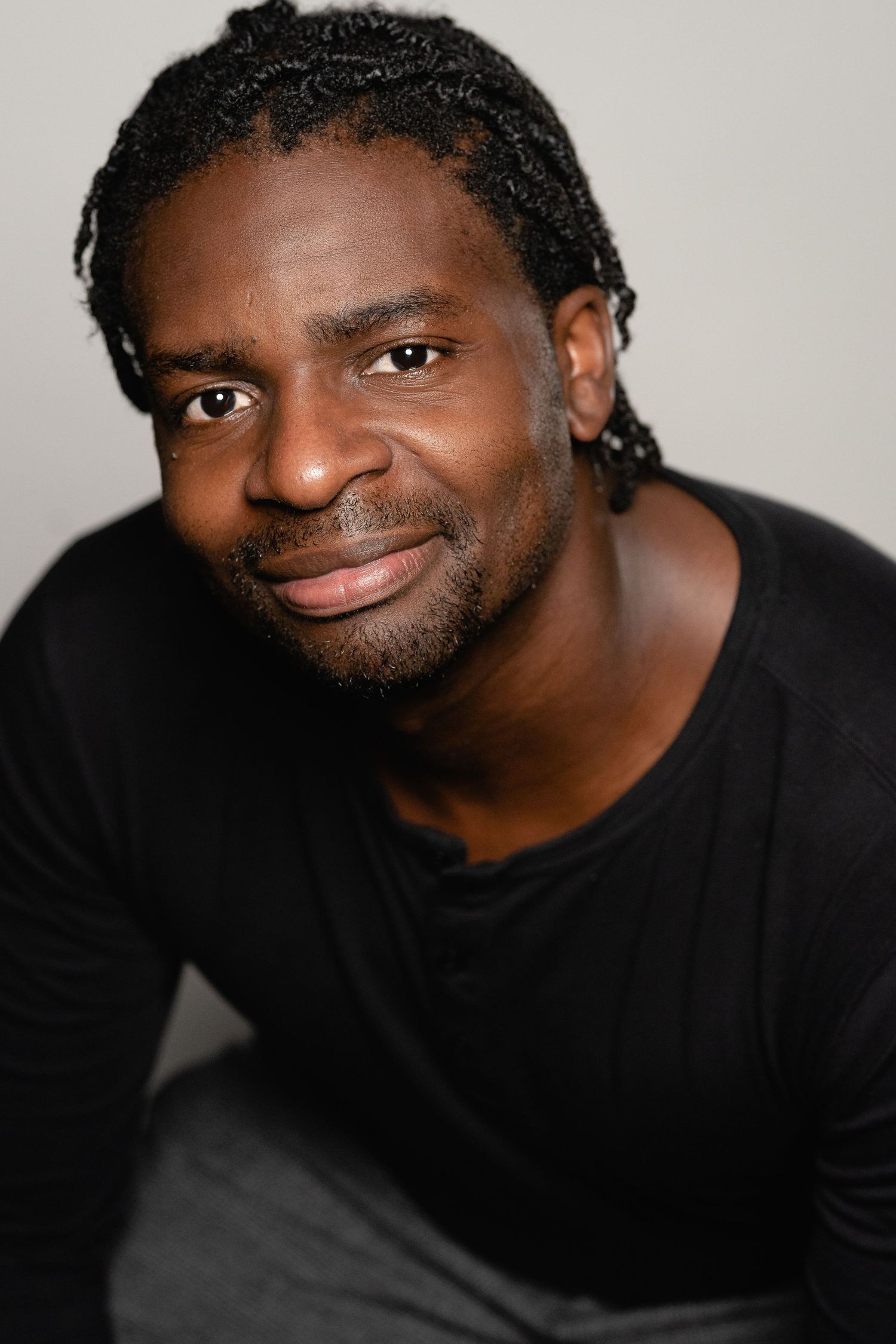 Smiling Black man with braided hair wearing a black shirt, portrait photo with neutral background.