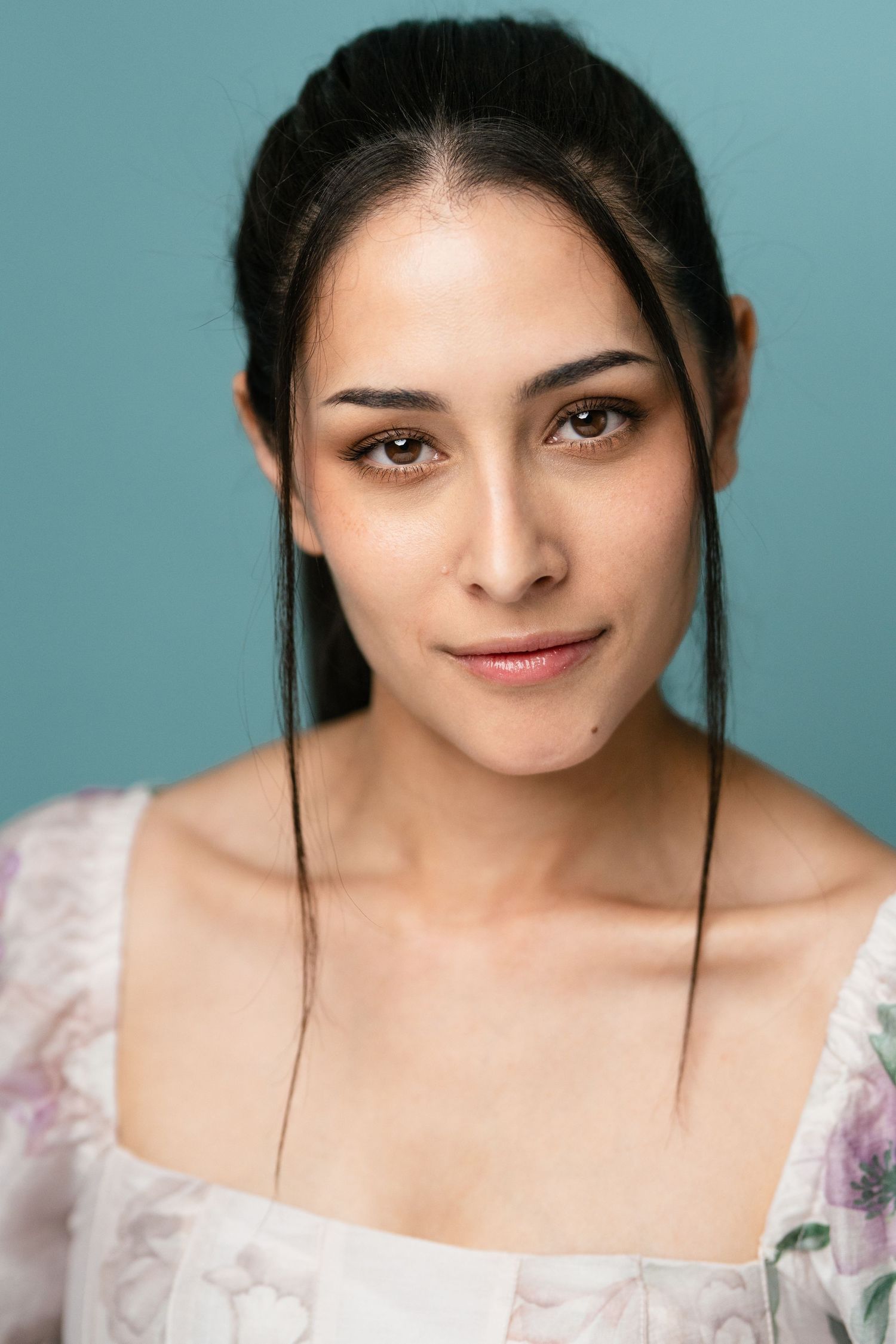 Young woman with dark hair in floral off-shoulder top poses against teal background in studio portrait.