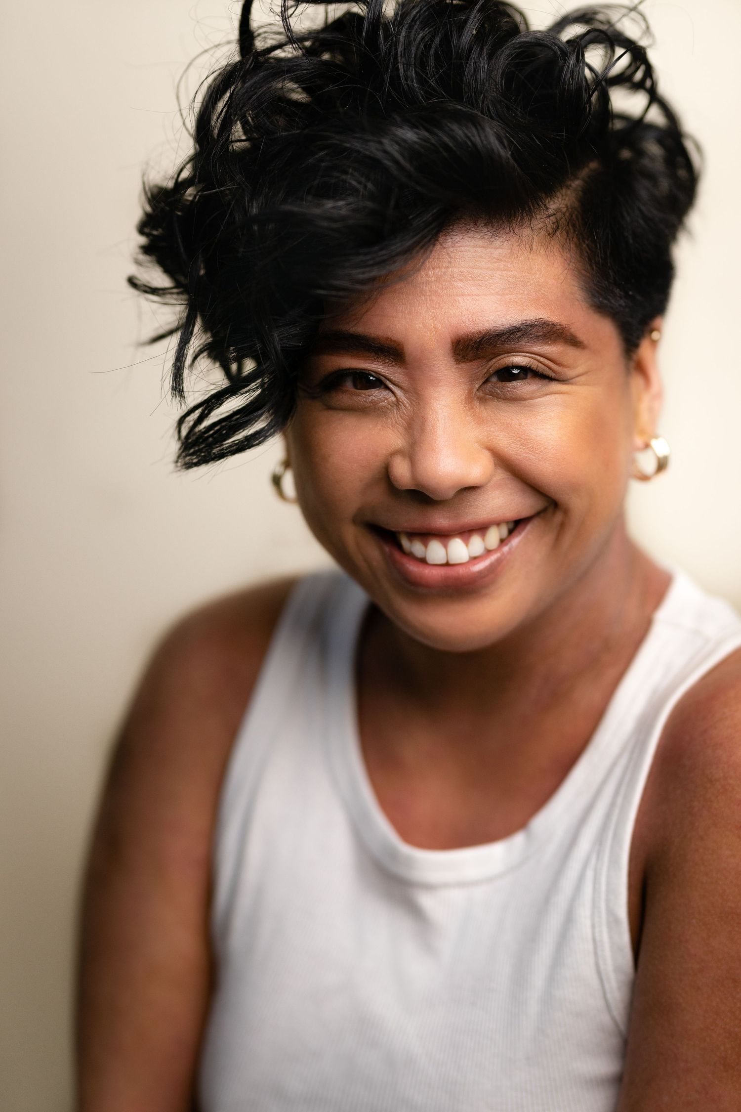 Smiling woman with short curly black hair wearing a white tank top and gold earrings against a light background.