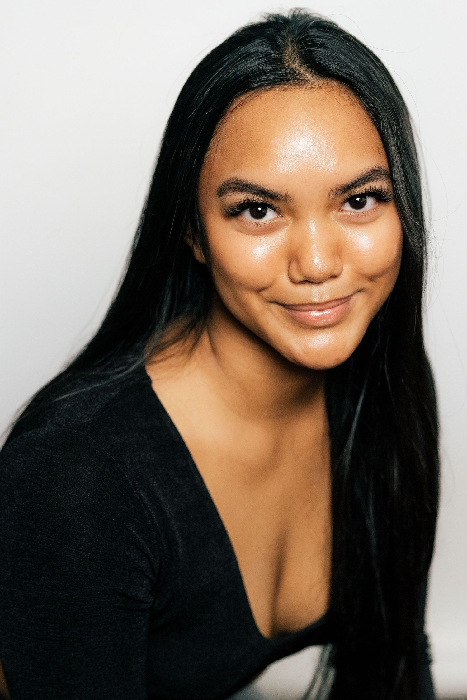 Young woman with long black hair and warm skin tone smiling softly, wearing a black top against a light background.