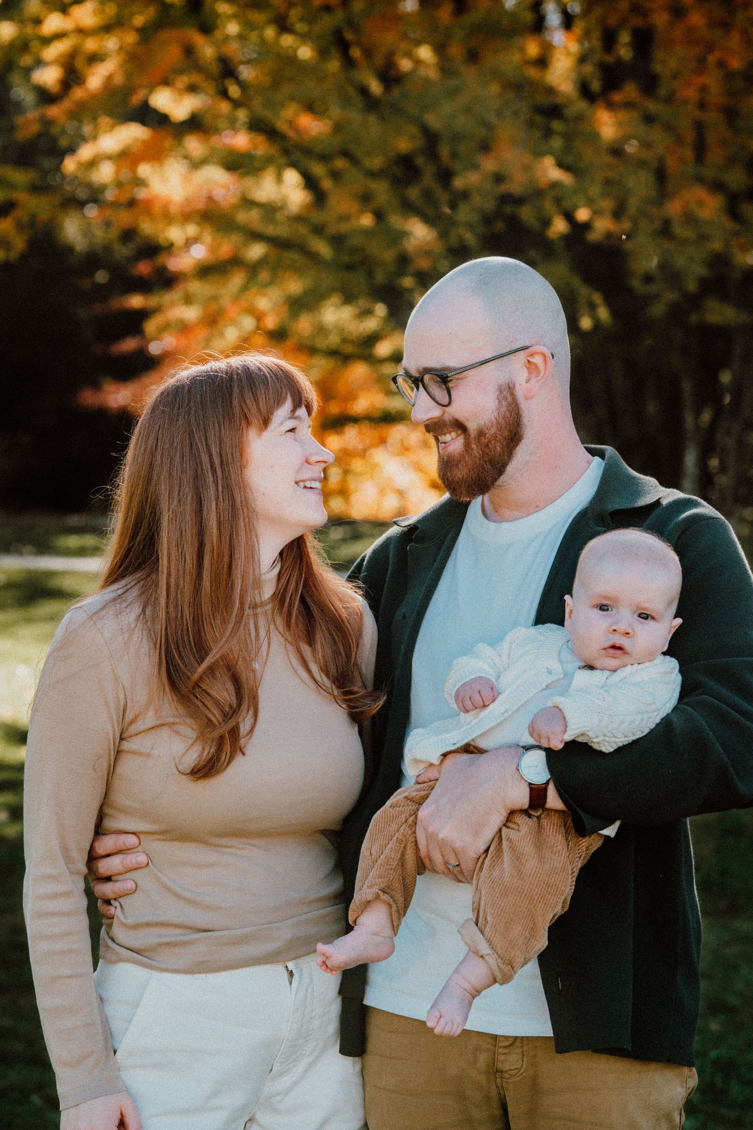 A smiling couple with a bald bearded man holding a baby outdoors among vibrant autumn foliage.