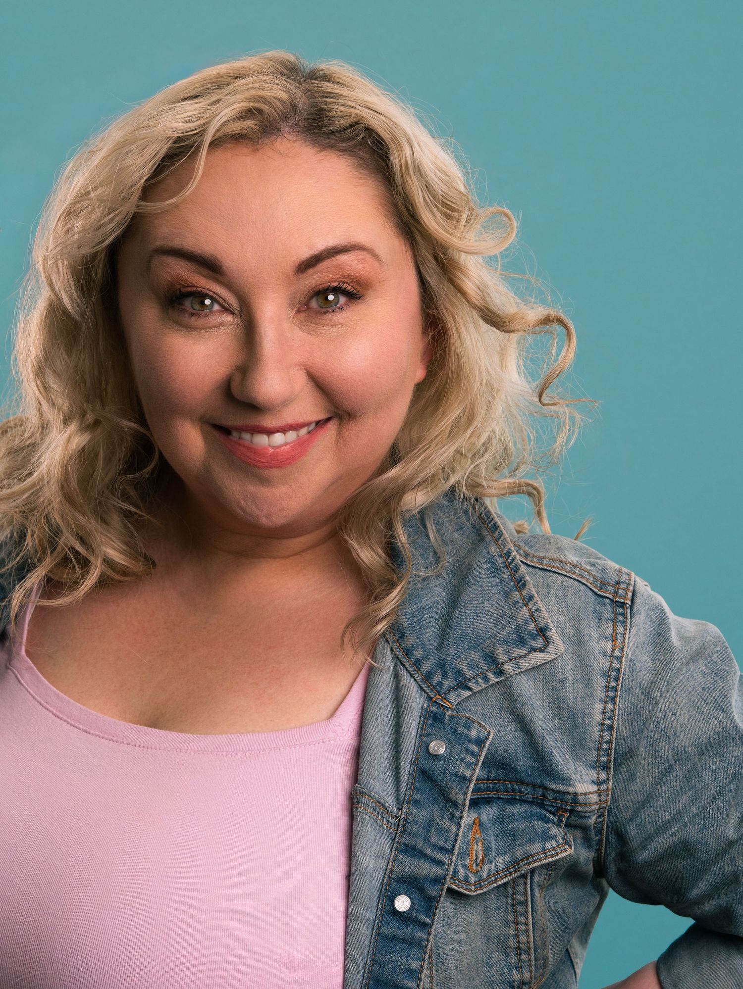 Smiling blonde woman in pink top and denim jacket poses against teal background.