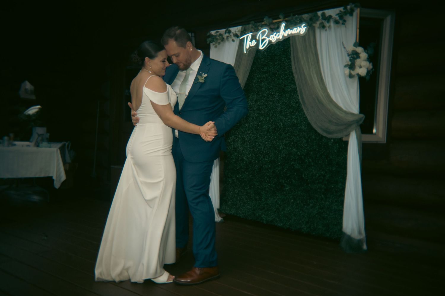 Bride and groom share first dance in front of green hedge wall with neon sign reading The Brehmes.