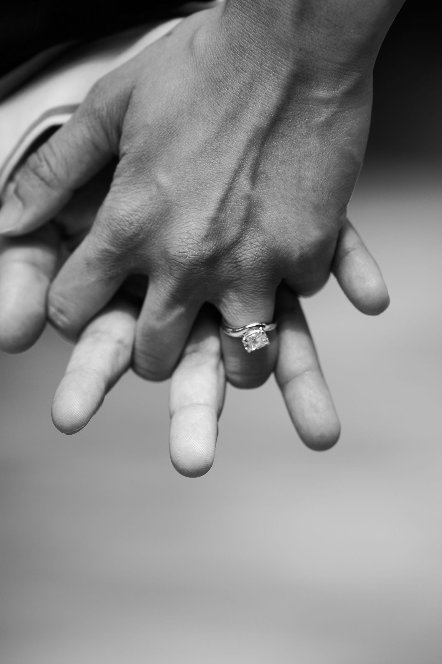 Black and white photo of two hands intertwined, showcasing a sparkling diamond engagement ring on the woman's finger.