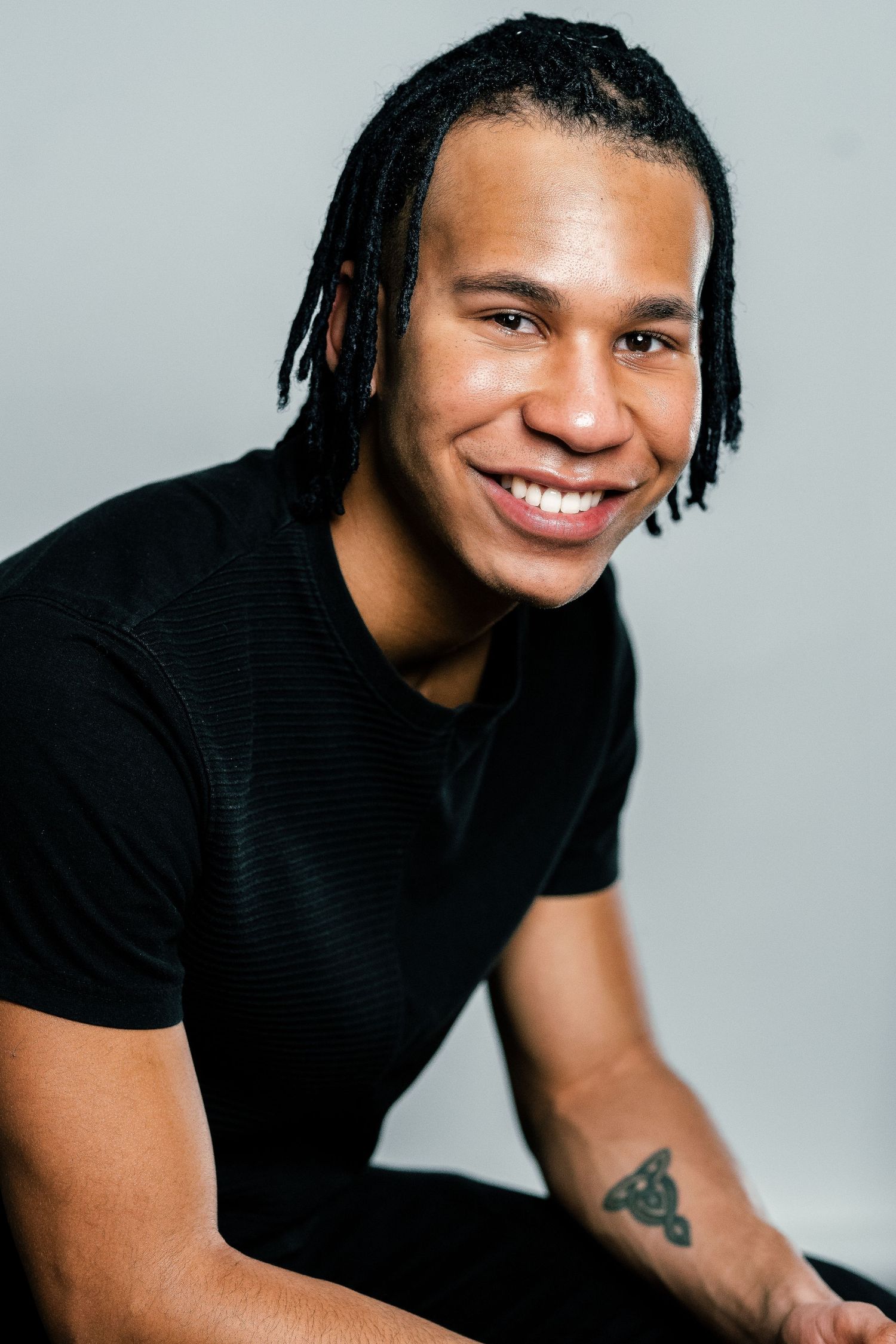 Smiling young man with dreadlocks and a tattoo on his forearm wearing a black t-shirt against a gray background.