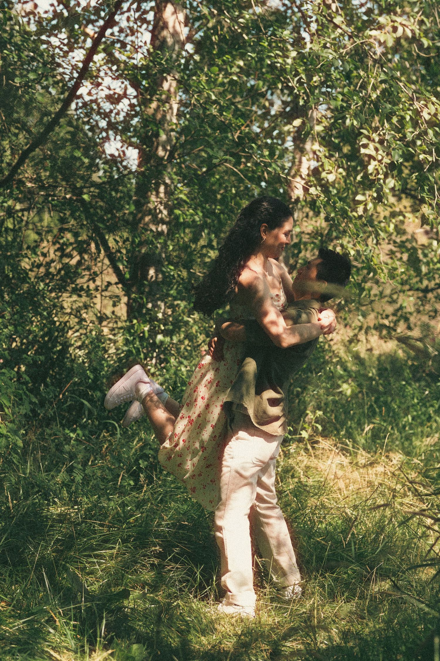 A couple embraces joyfully in a lush green forest, the woman lifted off the ground in a romantic outdoor setting.