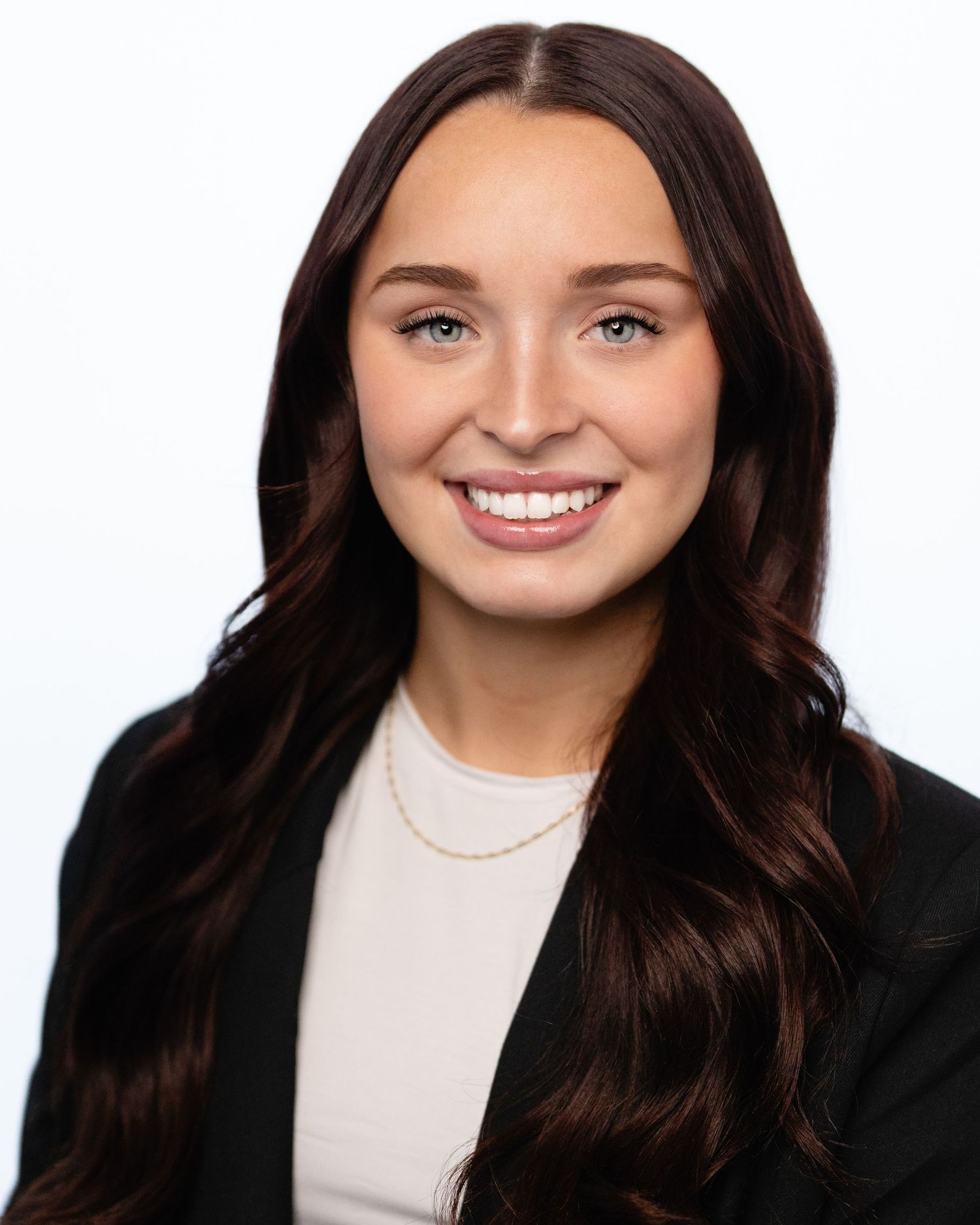 Professional headshot of a young woman with long dark hair, blue eyes, wearing a black blazer and gold necklace.