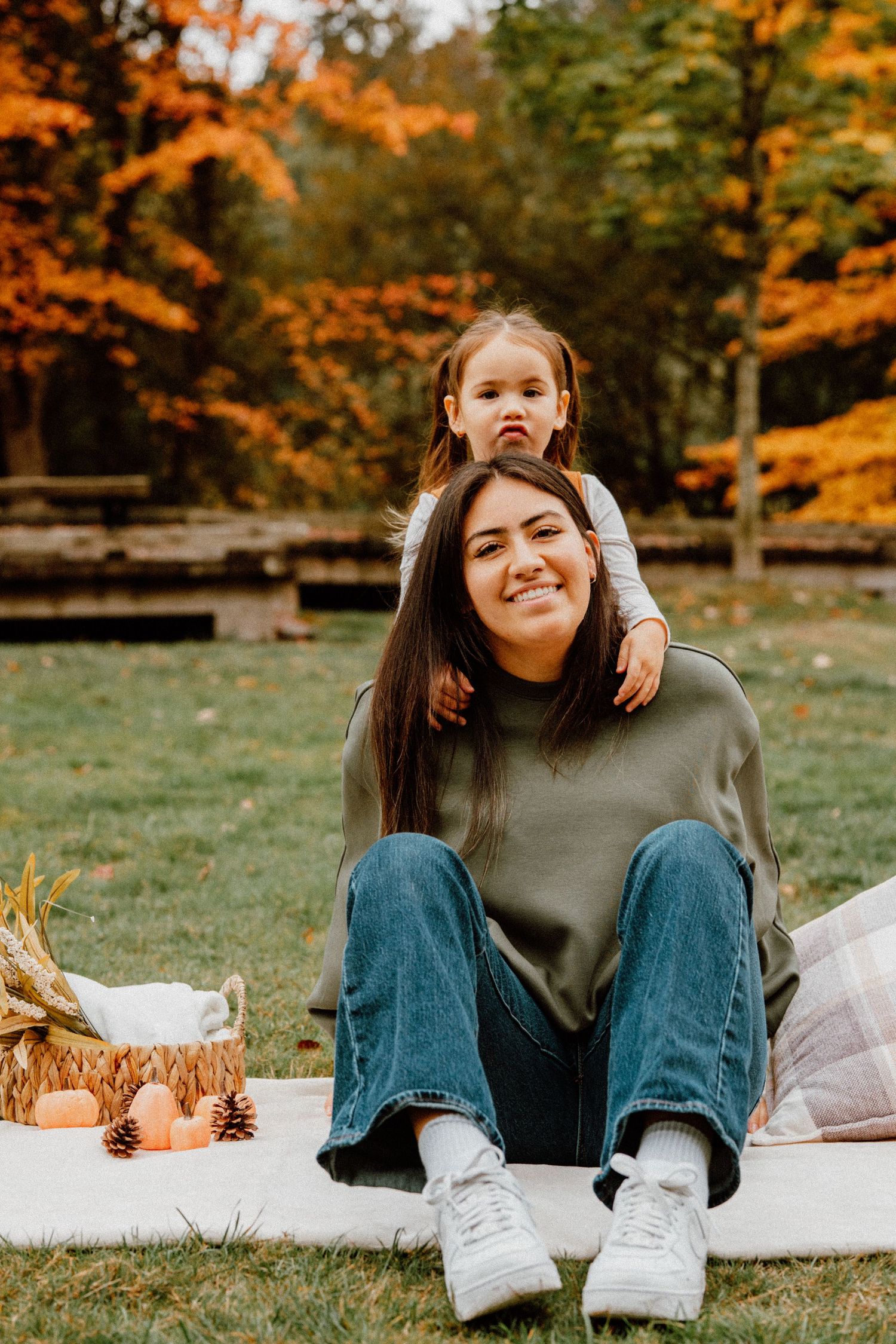 Woman and toddler daughter enjoying autumn picnic in colorful fall park, sitting on blanket with basket and pumpkins.