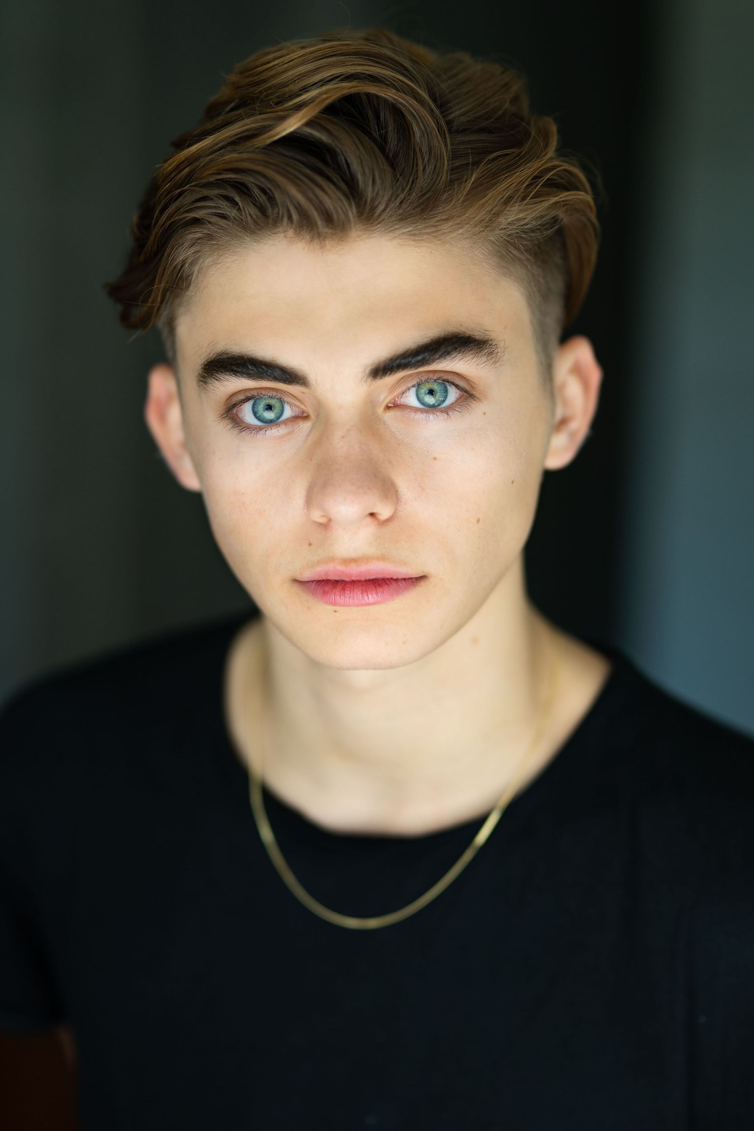 Young man with blue eyes, styled brown hair, gold chain necklace, wearing a black t-shirt, posing for a portrait photo.