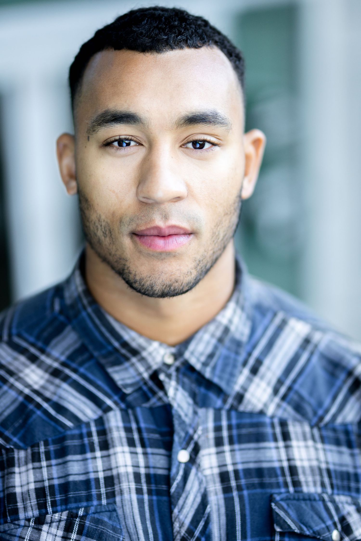 Young man in plaid shirt with short hair and light stubble poses for a professional headshot outdoors.