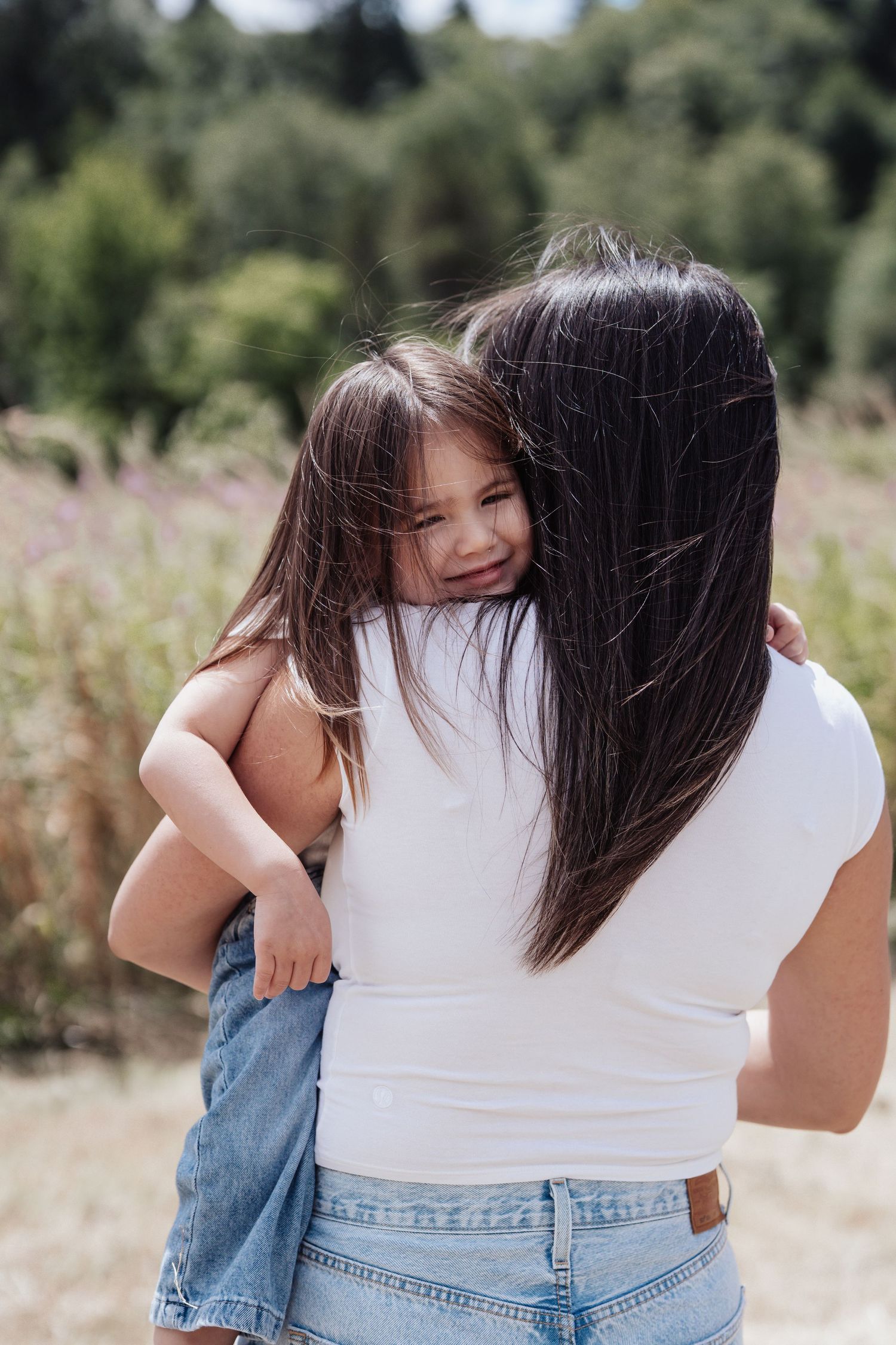 A mother in a white t-shirt and jeans holds her smiling young daughter outdoors in a field.