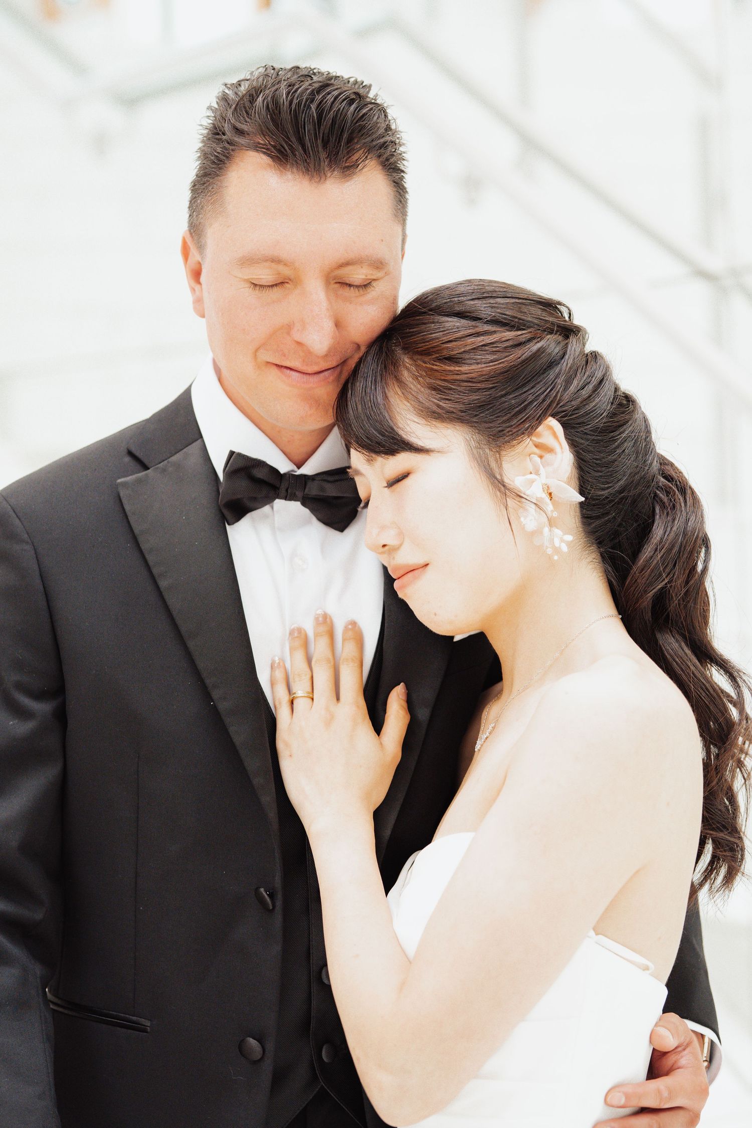 A bride and groom in formal wedding attire share a tender moment, with the bride resting her head on the groom's chest.