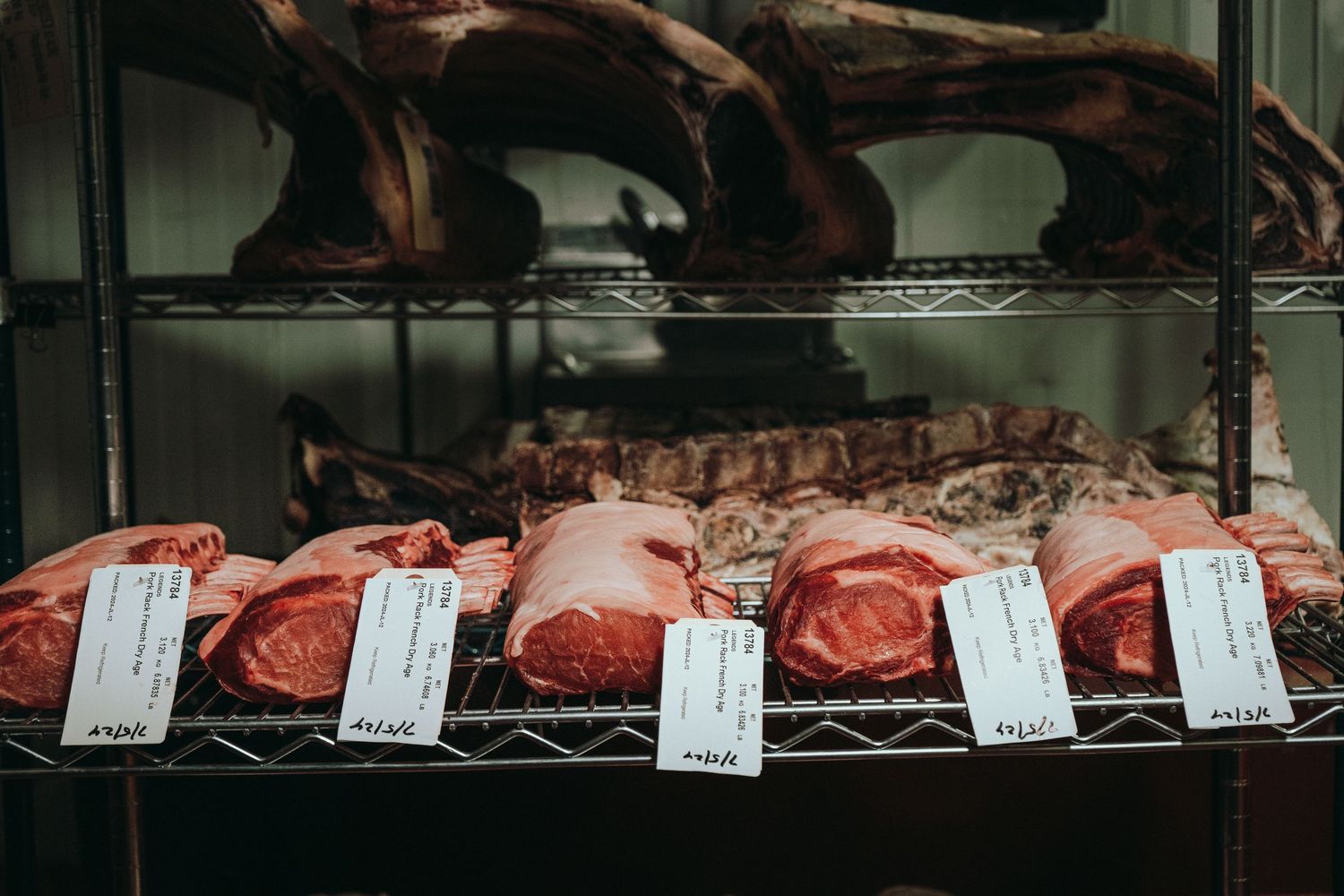 Assorted cuts of fresh raw meat displayed on metal shelves with price tags in a butcher shop.