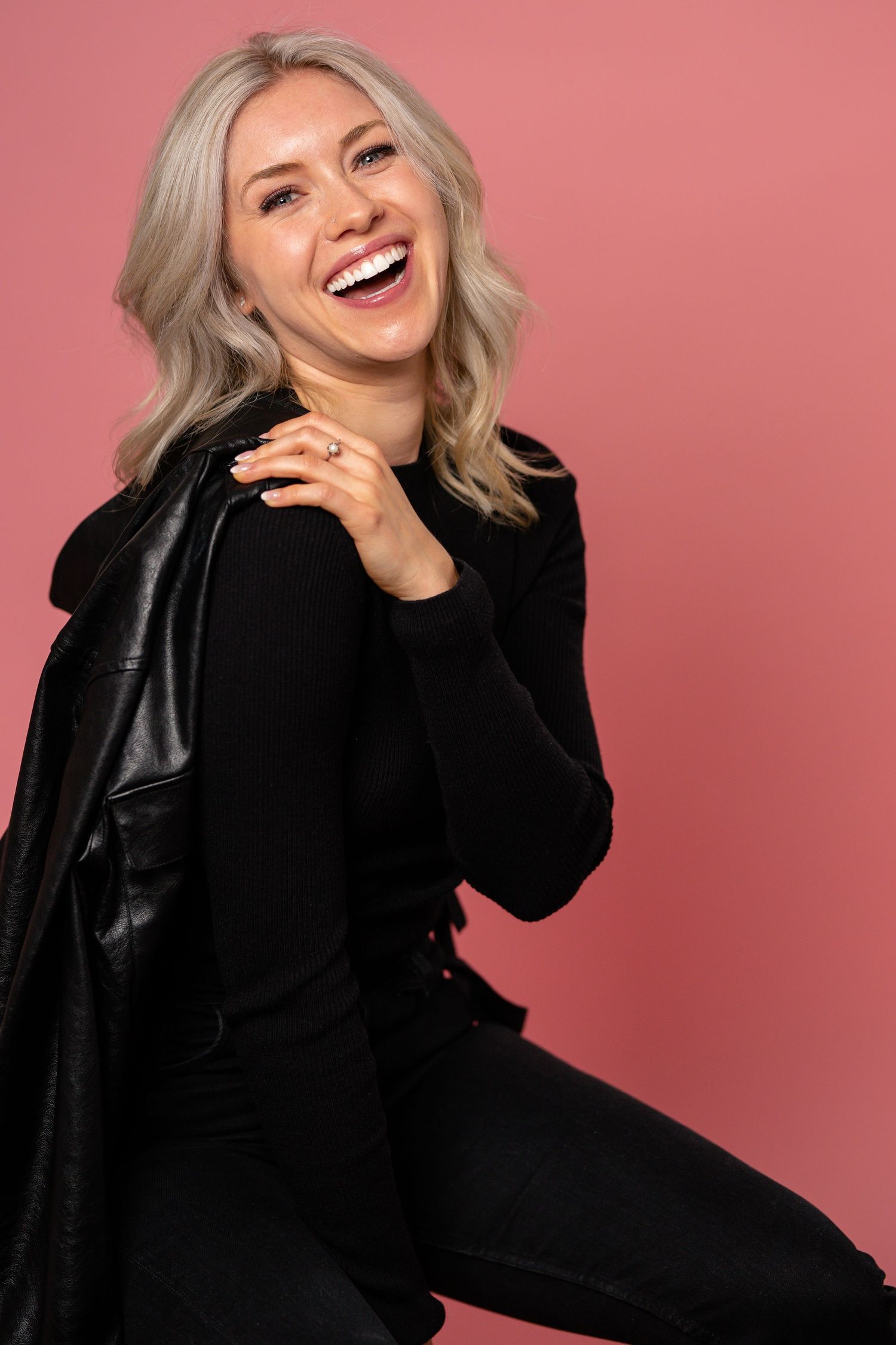 Laughing blonde woman in black outfit and leather jacket posing against a pink background.