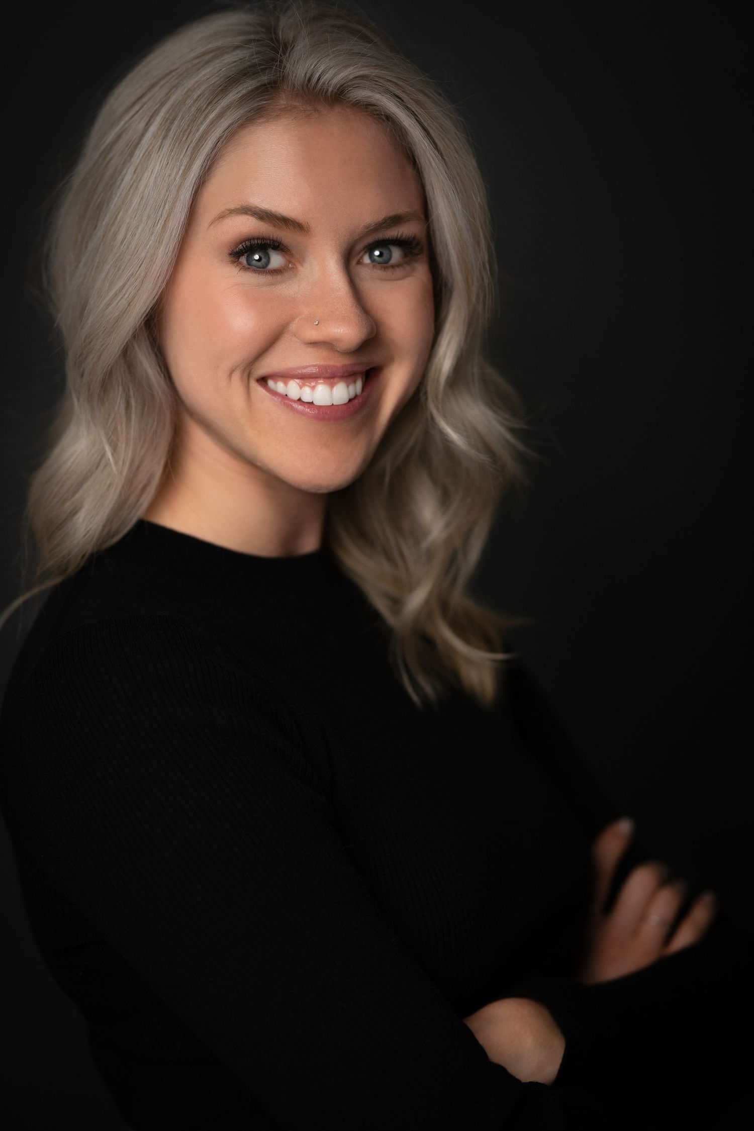 A smiling woman with blonde wavy hair wearing a black top, arms crossed, against a dark background.
