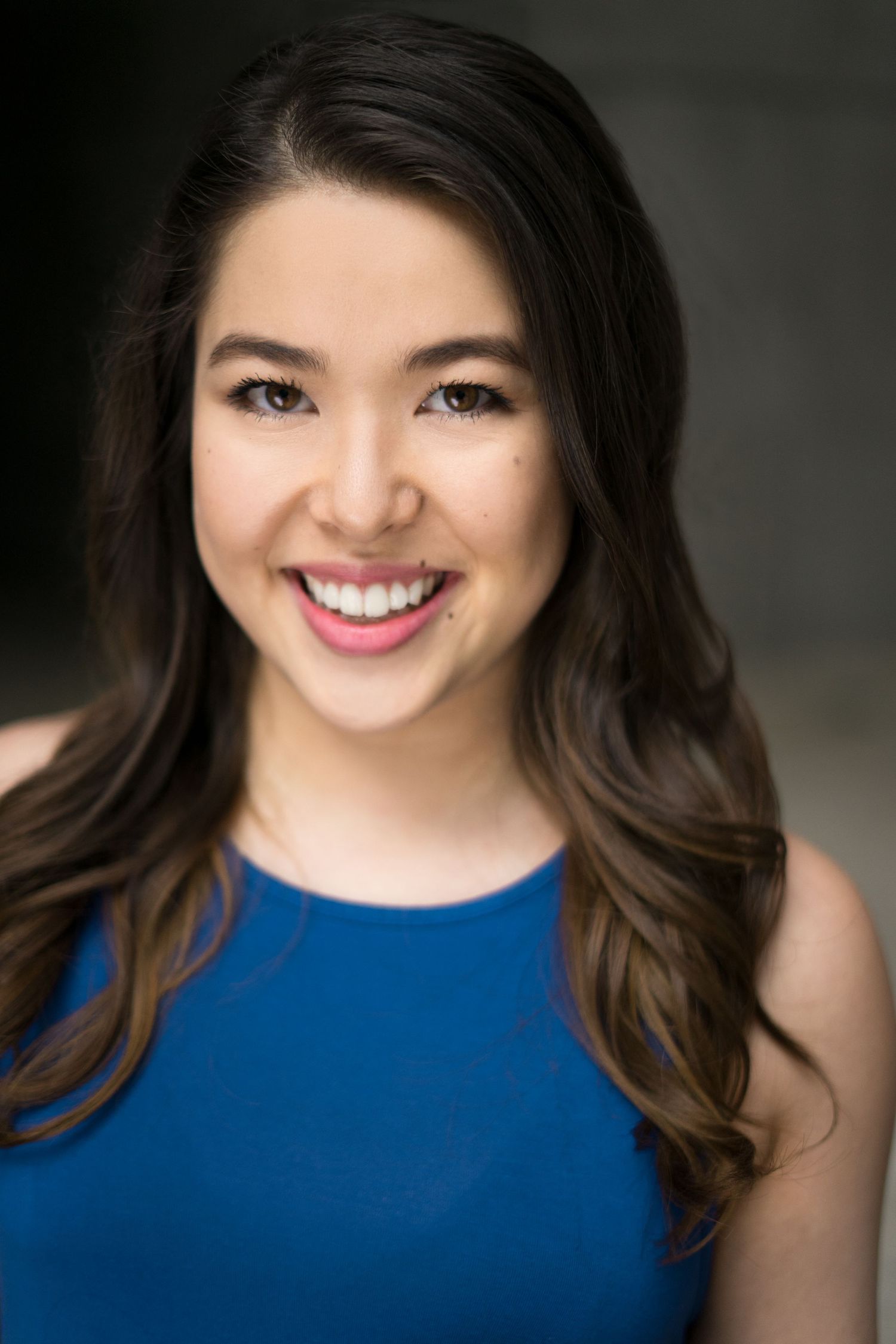 Young woman with long brown hair smiling in a blue top, professional headshot portrait.