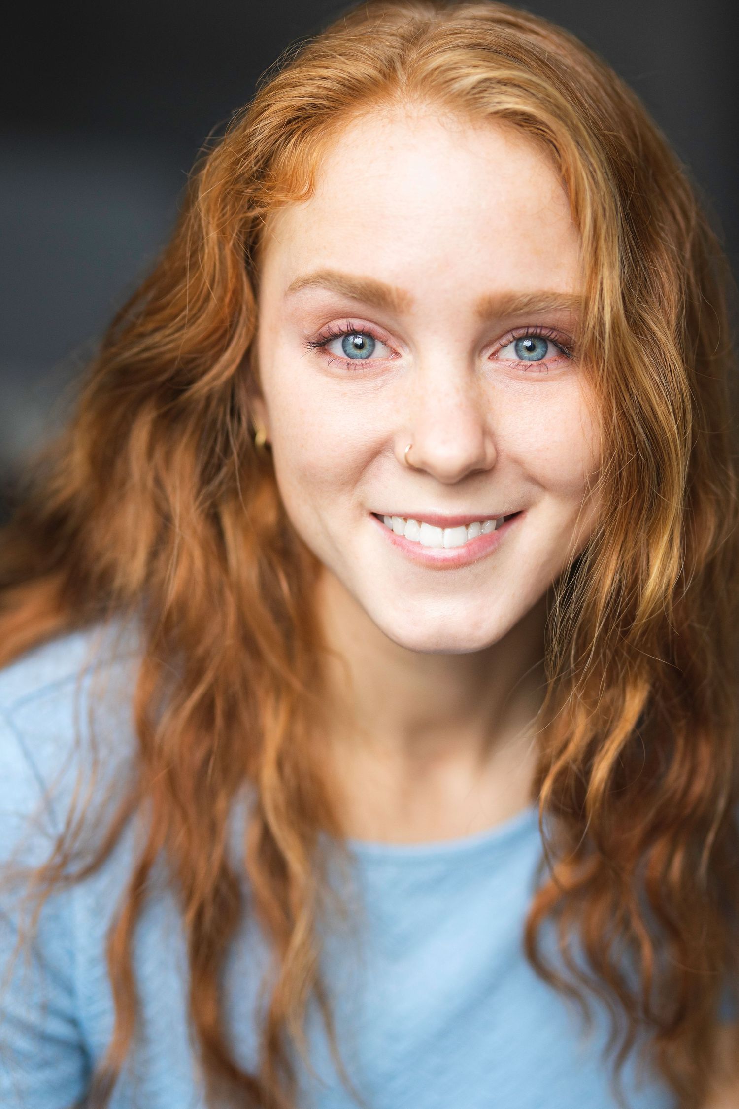 Smiling young woman with long wavy red hair, blue eyes, and freckles wearing a light blue top.