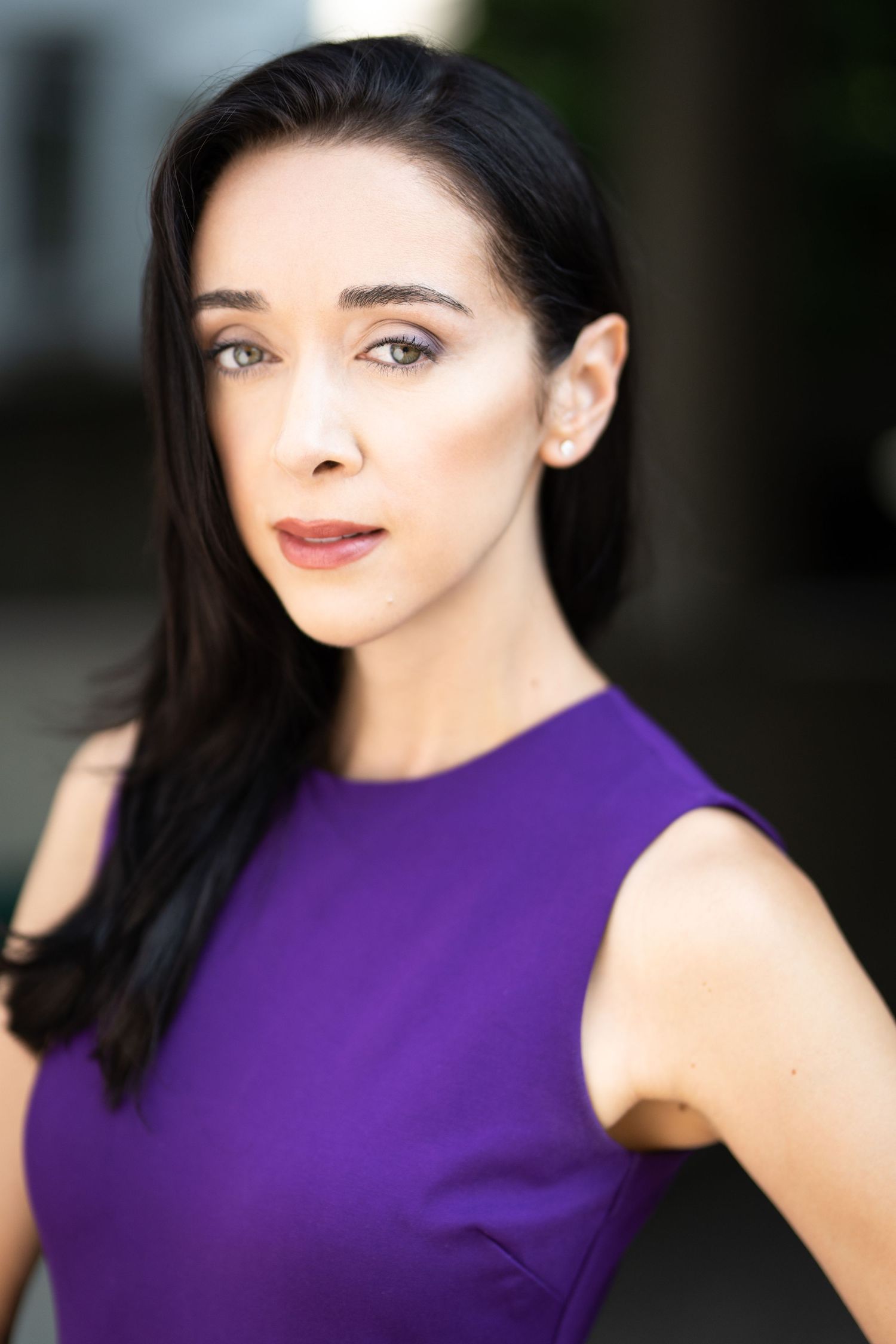 A woman with dark hair wearing a purple sleeveless dress poses for a professional headshot outdoors.