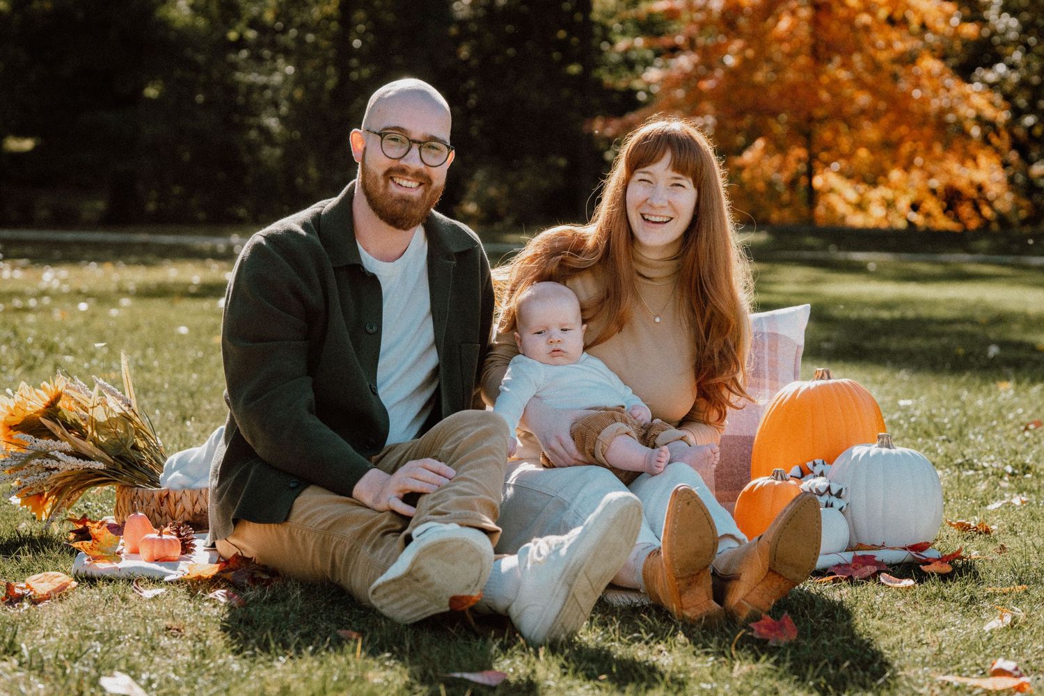 Family of three sitting on grass in autumn park with pumpkins, parents smiling holding baby during fall photo session.