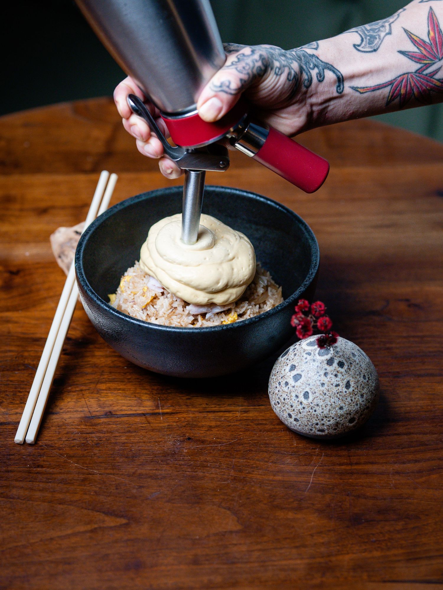 Tattooed hand using a meat press on fried rice in a black bowl, with chopsticks and decorative stone sphere on wooden table.