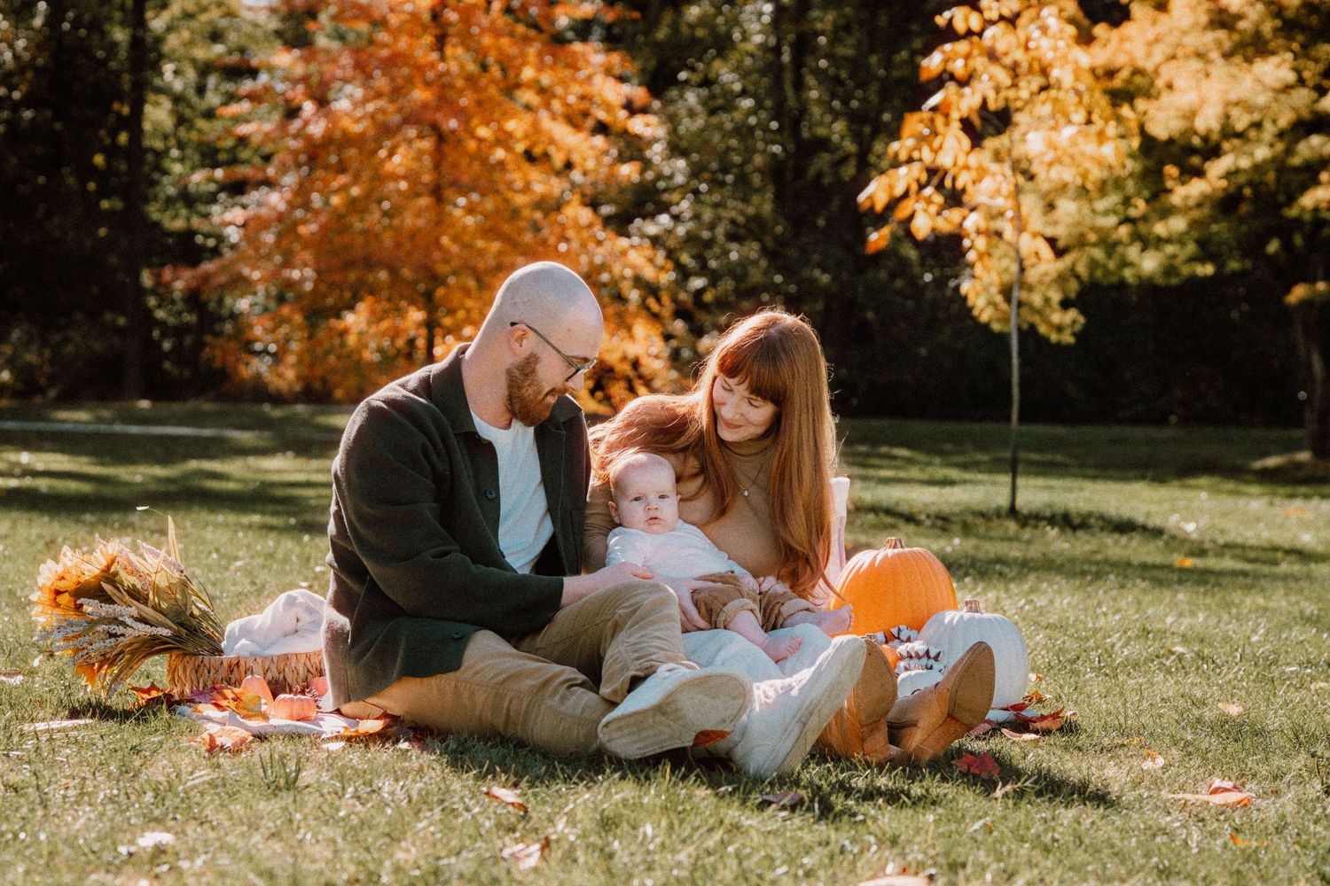 Family sitting on a blanket in a sunny autumn park with orange pumpkins and colorful fall foliage.