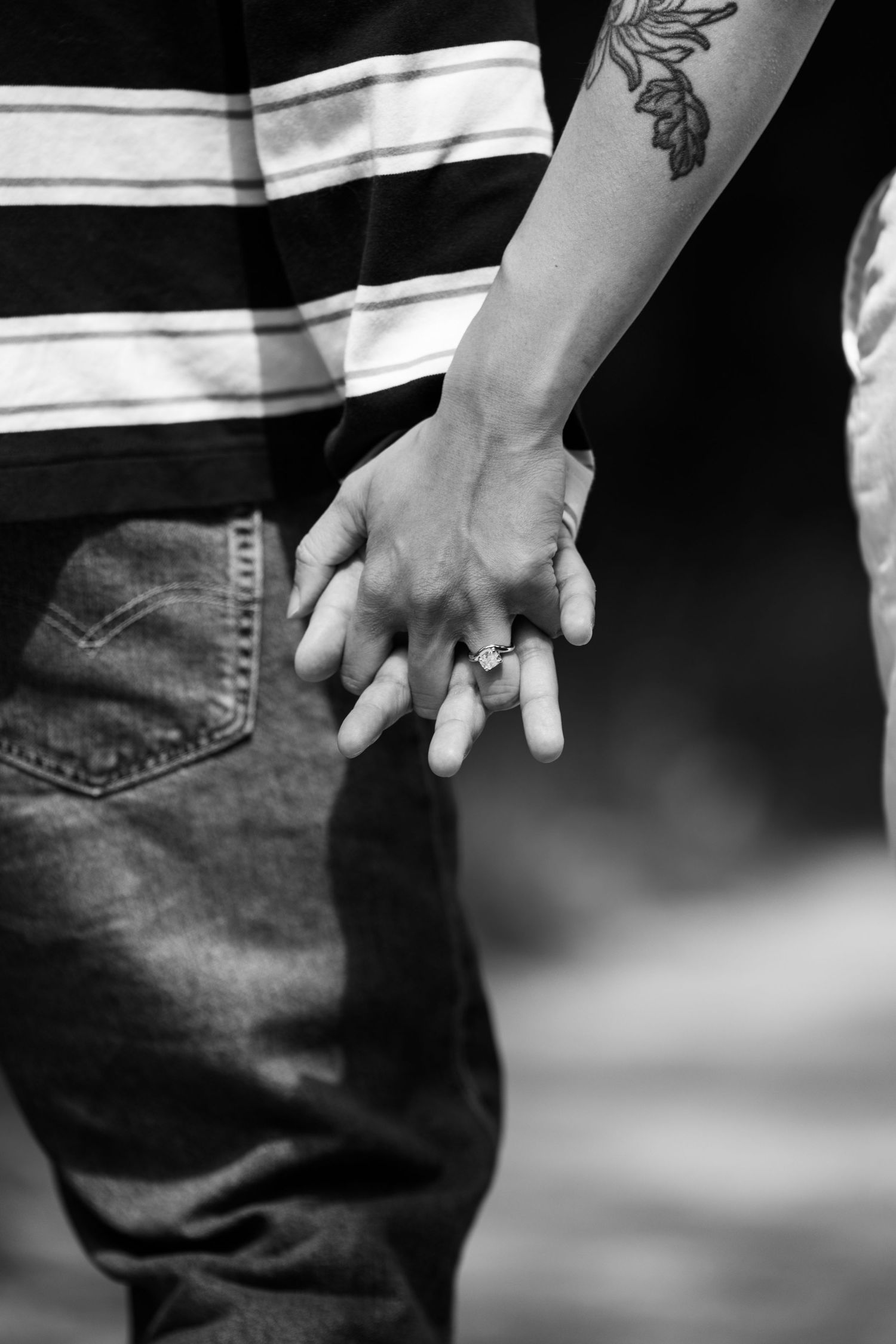 Black and white photo of a couple holding hands, with a floral tattoo and engagement ring visible.