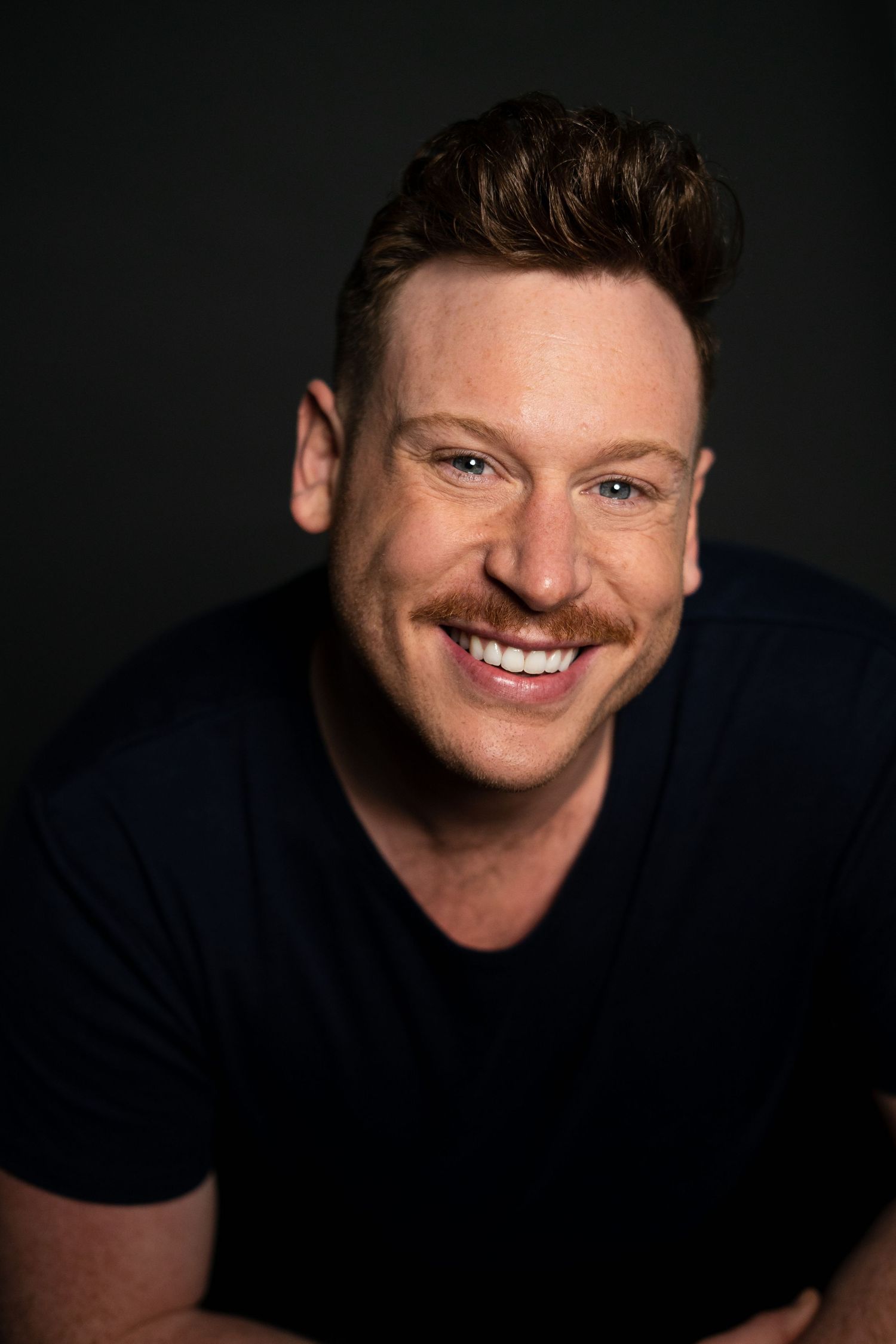 Smiling man with auburn hair and mustache wearing a navy blue t-shirt against a dark background.