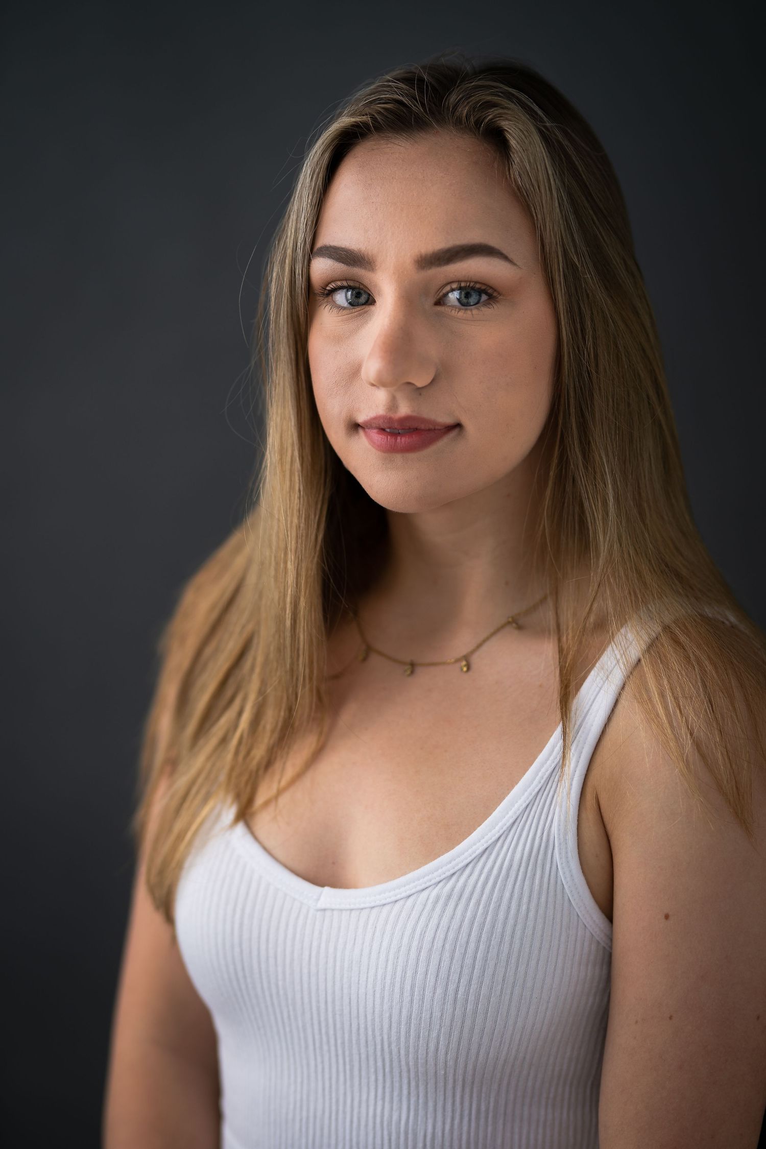 Young blonde woman in white ribbed tank top poses against dark gray background, wearing delicate gold necklace.