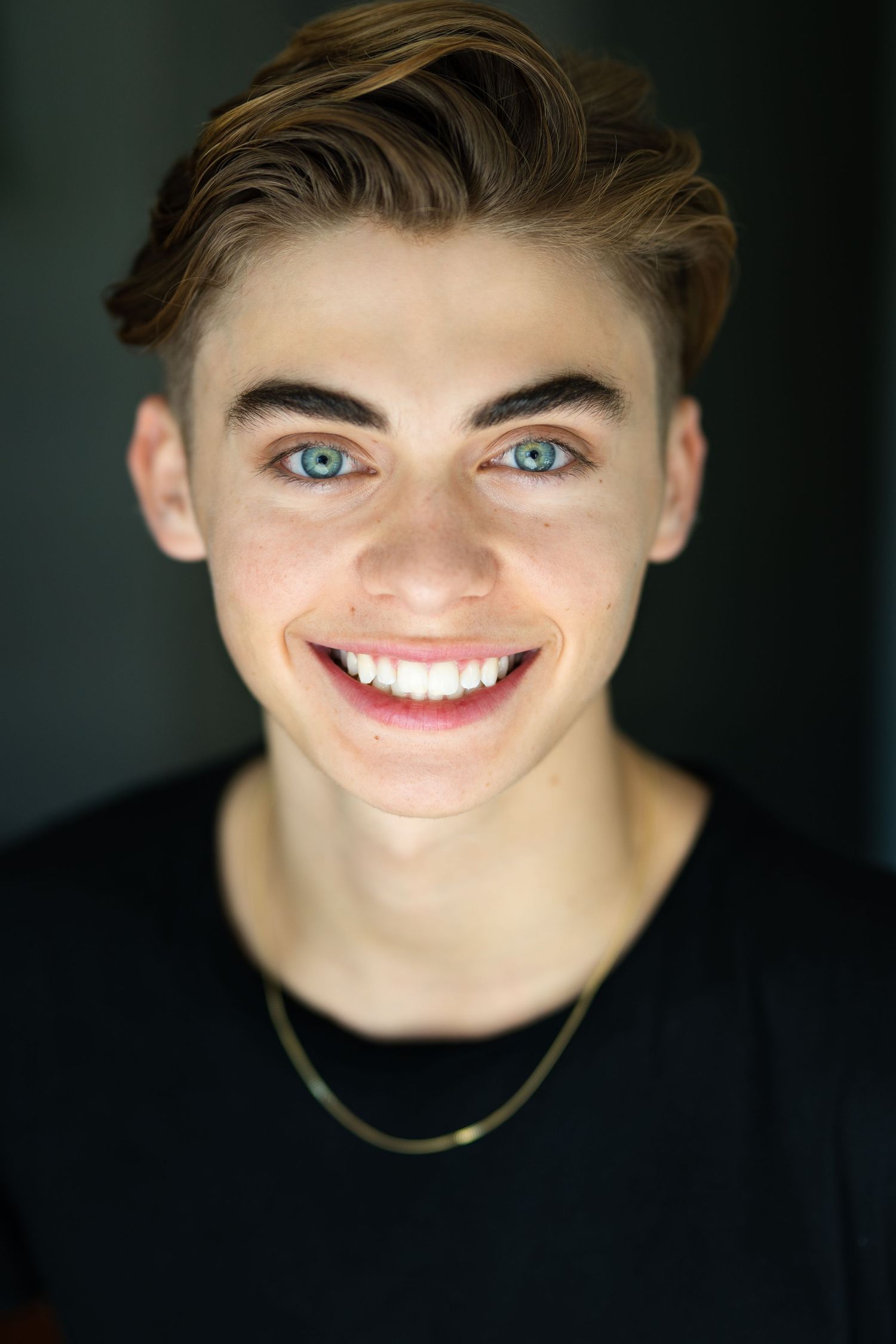 Smiling young man with blue eyes, styled hair, and a gold chain necklace wearing a black shirt.