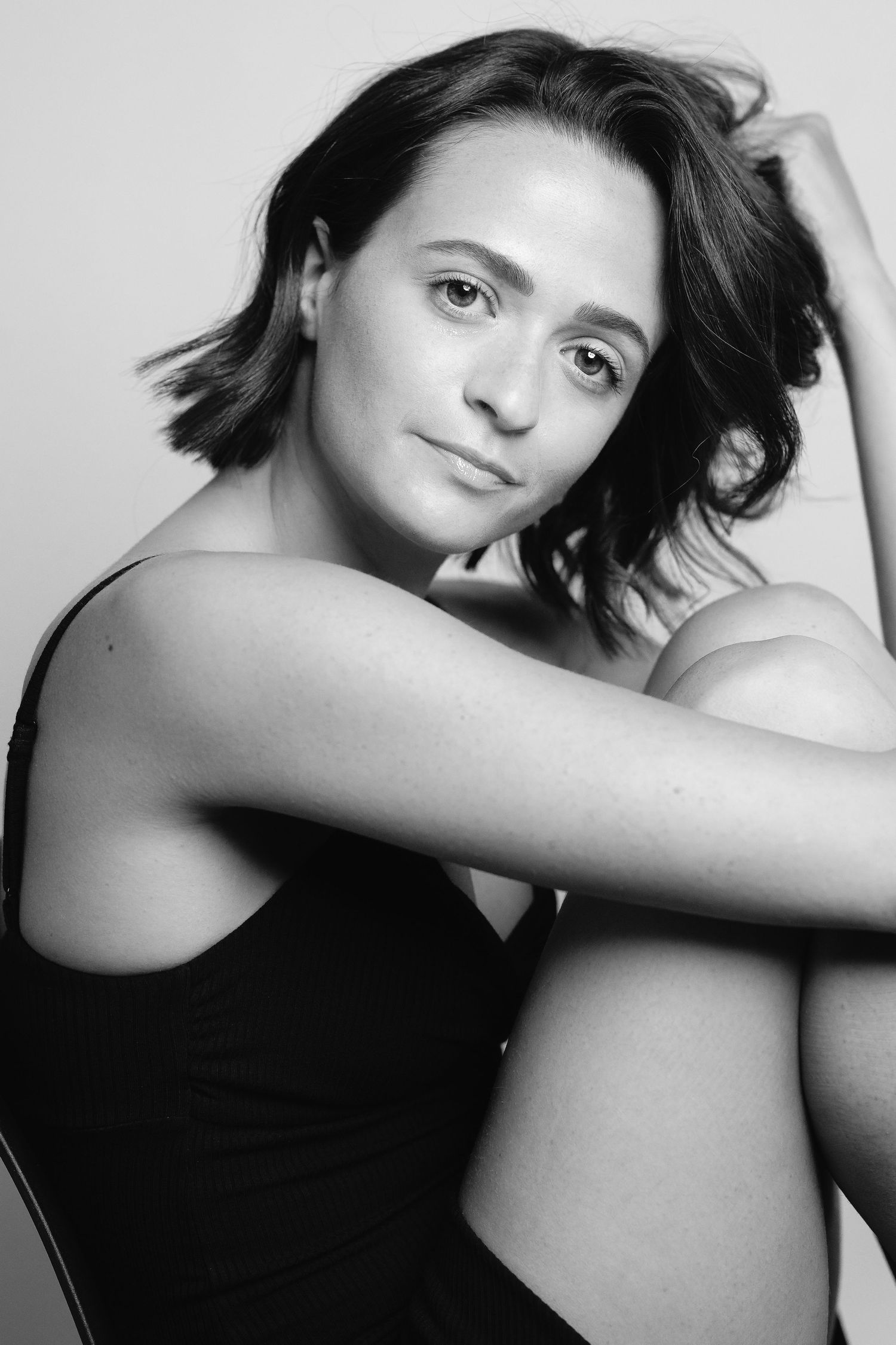 Black and white portrait of a young woman with short dark hair, sitting with arms crossed over knees, looking at camera.
