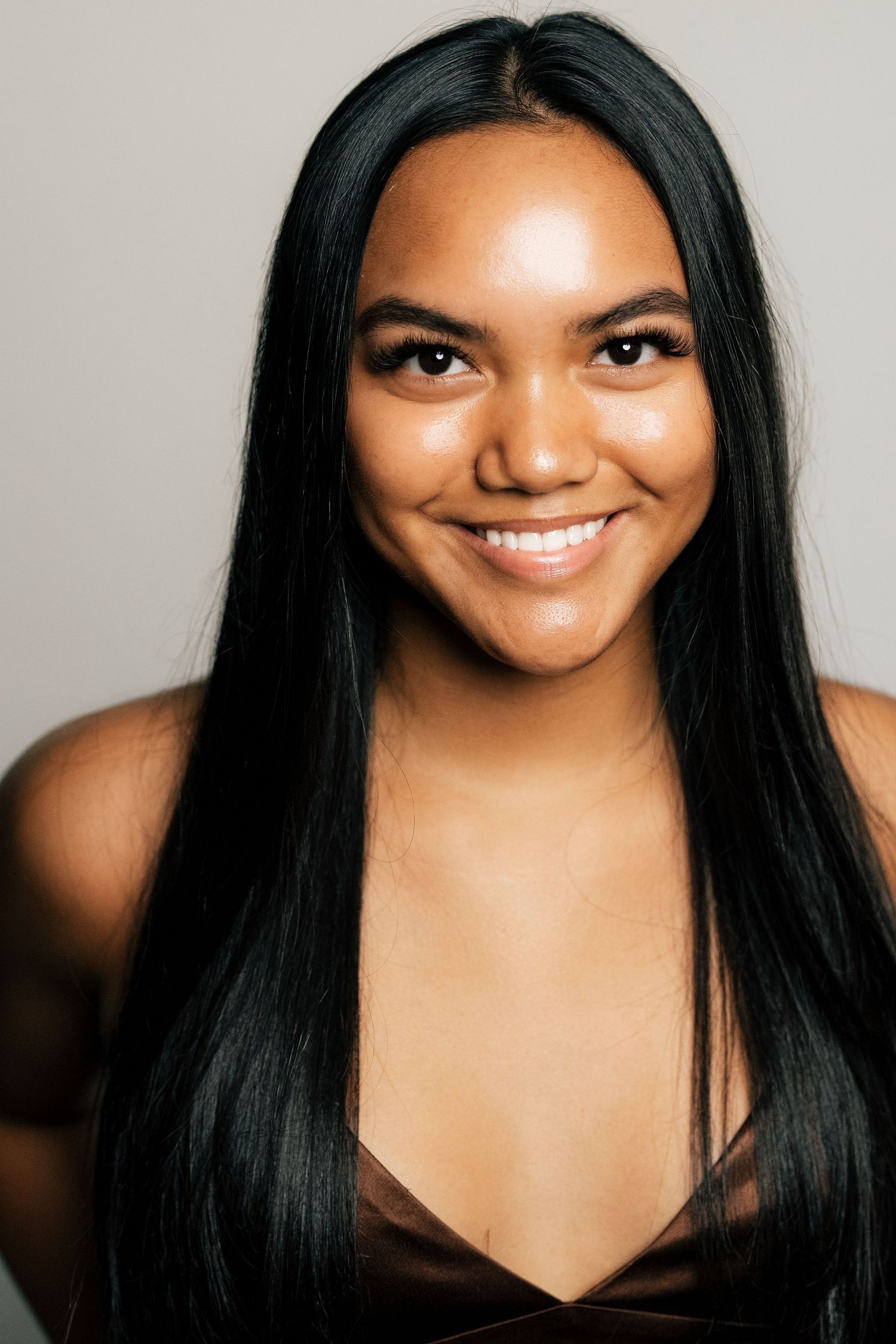 Young woman with long black hair smiling confidently, wearing a brown top against a neutral gray background.