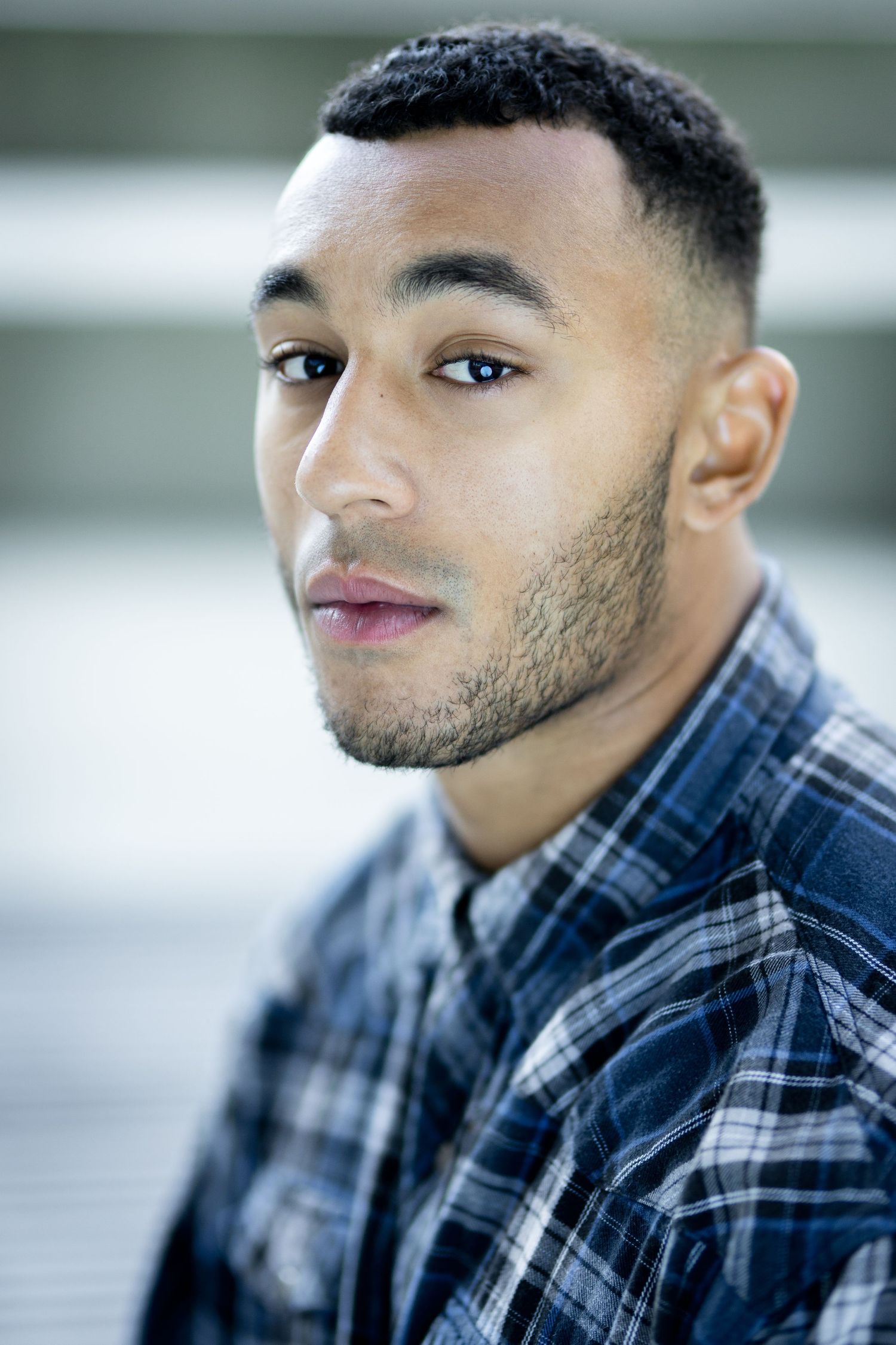 Young man with short hair and beard wearing a plaid shirt, looking at camera with a neutral expression.