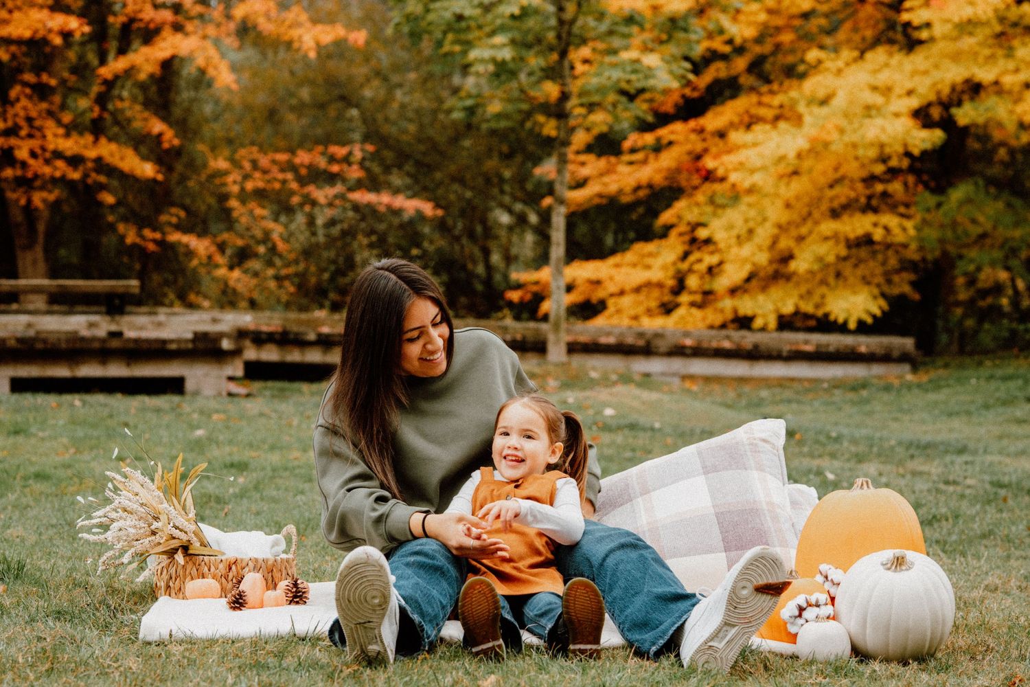 Mother and toddler laughing together on a blanket surrounded by fall pumpkins and colorful autumn trees.