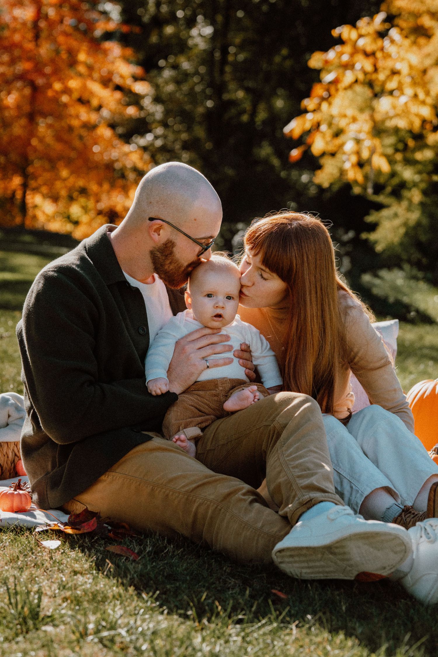 A couple sits on grass in autumn park, kissing their baby, surrounded by golden foliage in warm sunlight.