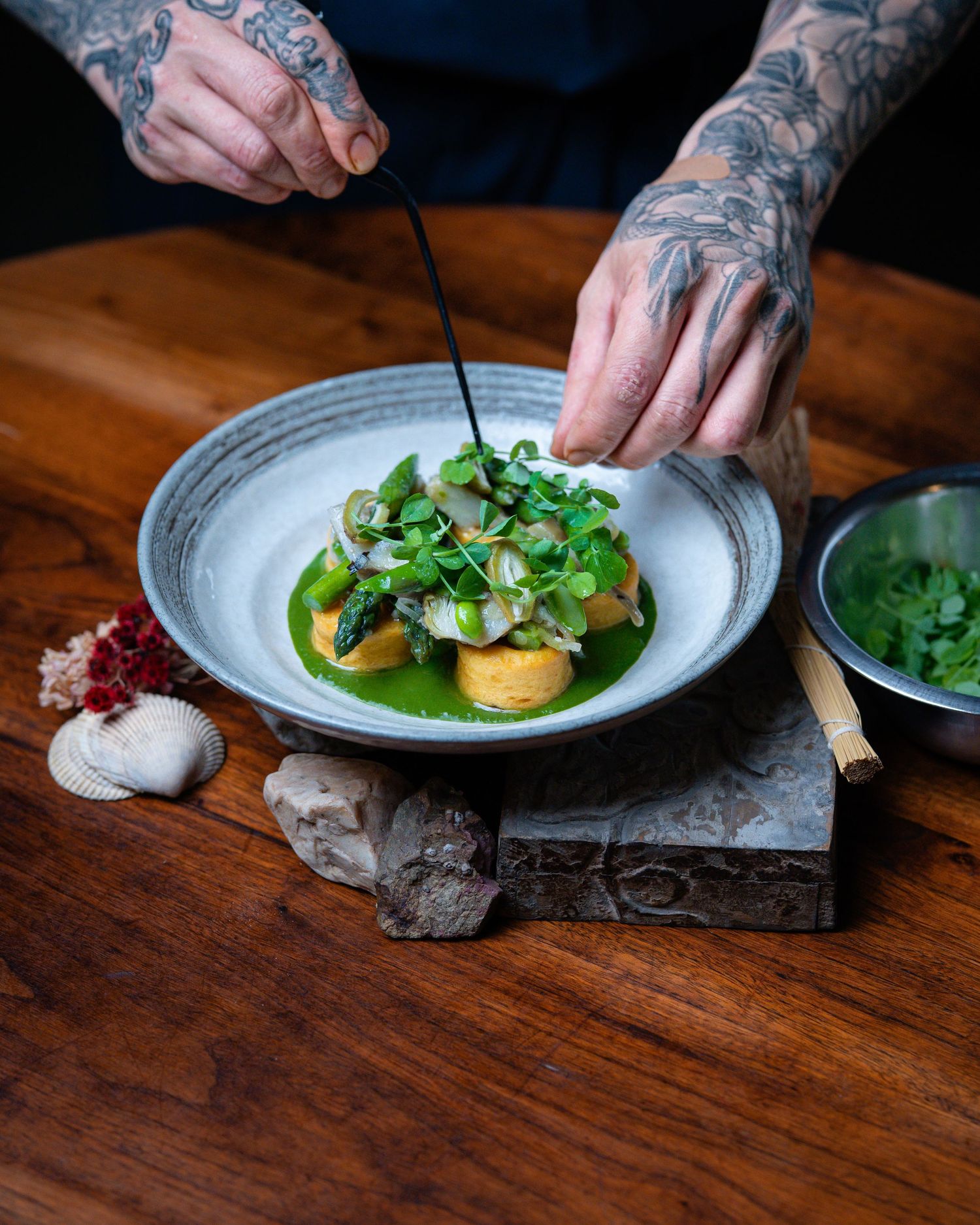 Tattooed chef's hands garnishing a vibrant green herb sauce dish with microgreens on a rustic wooden table.
