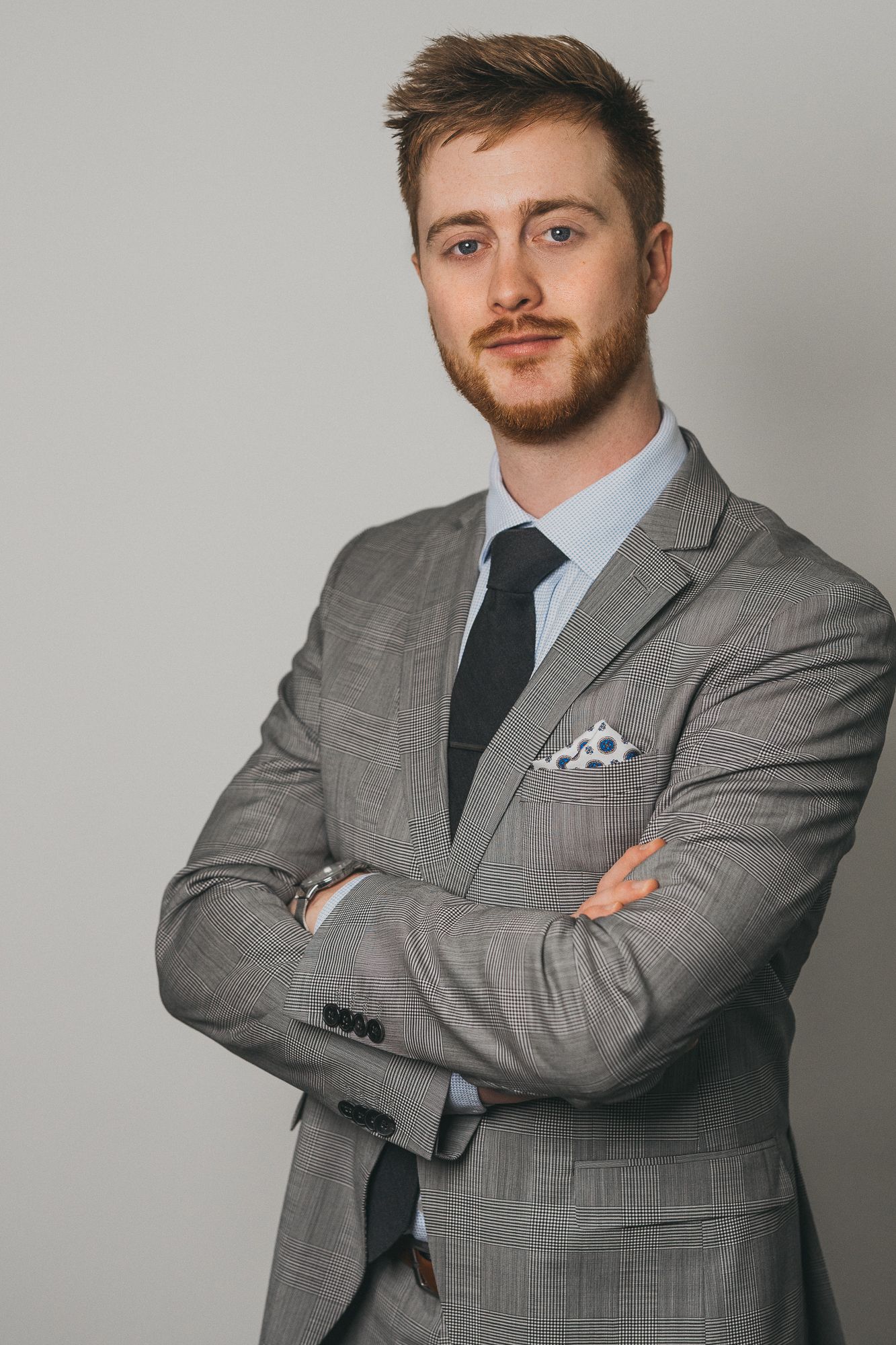 Young professional man in gray suit with crossed arms, dark tie, and pocket square poses confidently against light background.